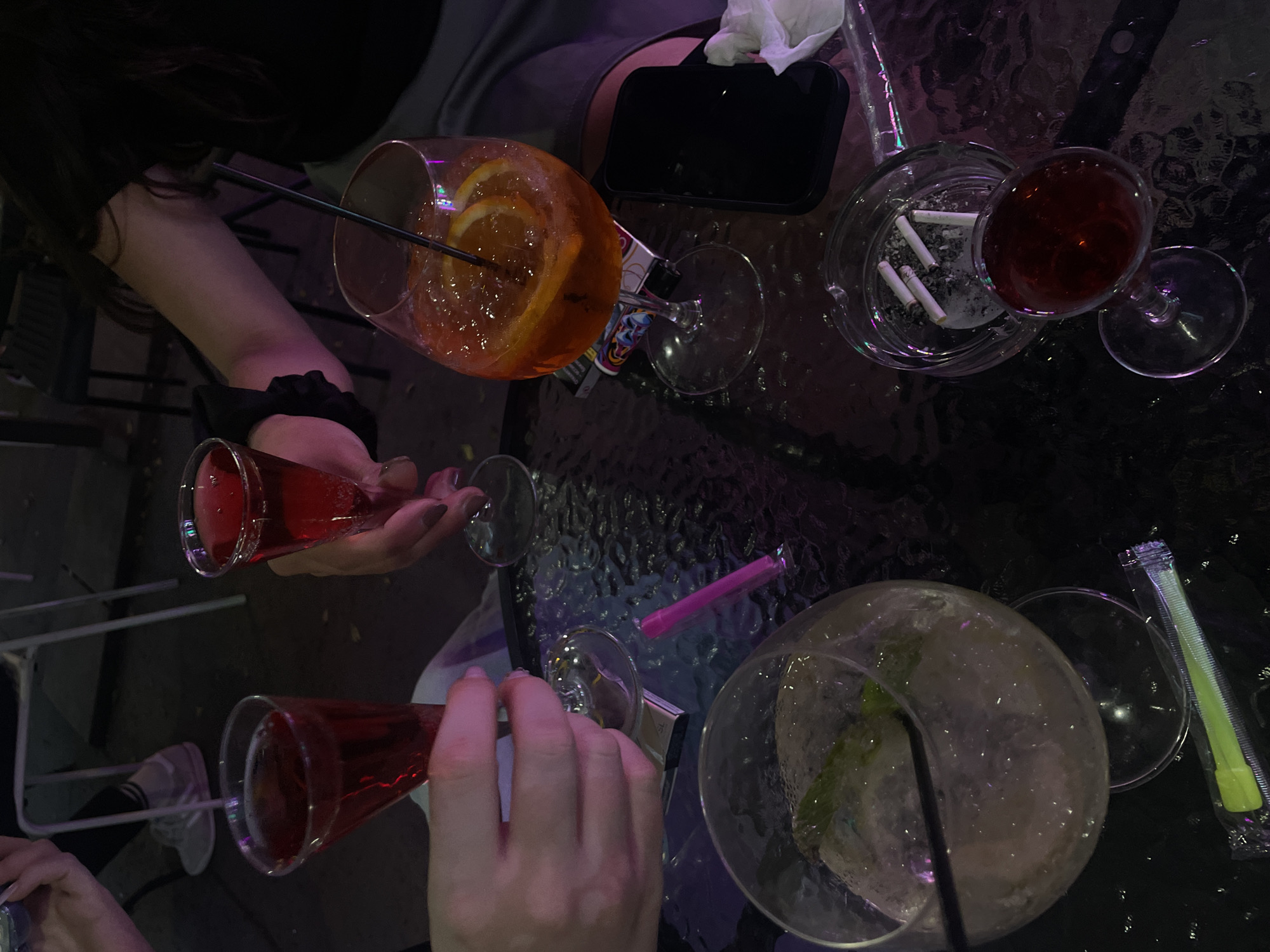 The image depicts an outdoor seating area at night, likely a cafe or bar in Budapest, Hungary. A dark, glass-topped table is laden with various items. In the foreground, a round, clear glass contains ice and a pale beverage with mint leaves, accompanied by a black straw and a packaged neon green straw. To the right, an ashtray holds several cigarette butts, and a small stemmed glass contains a dark red liquid. Further back, an Aperol Spritz is served in a wine glass with orange slices and a black straw. Two other glasses, one tall and slender, containing a red beverage, are being held by two individuals whose hands and arms are visible. One of the individuals is holding a lighter. A black smartphone is also visible on the table. In the background, the legs and feet of individuals are seen, some wearing sneakers, and outdoor chairs with metal frames. The overall ambiance suggests a social gathering.