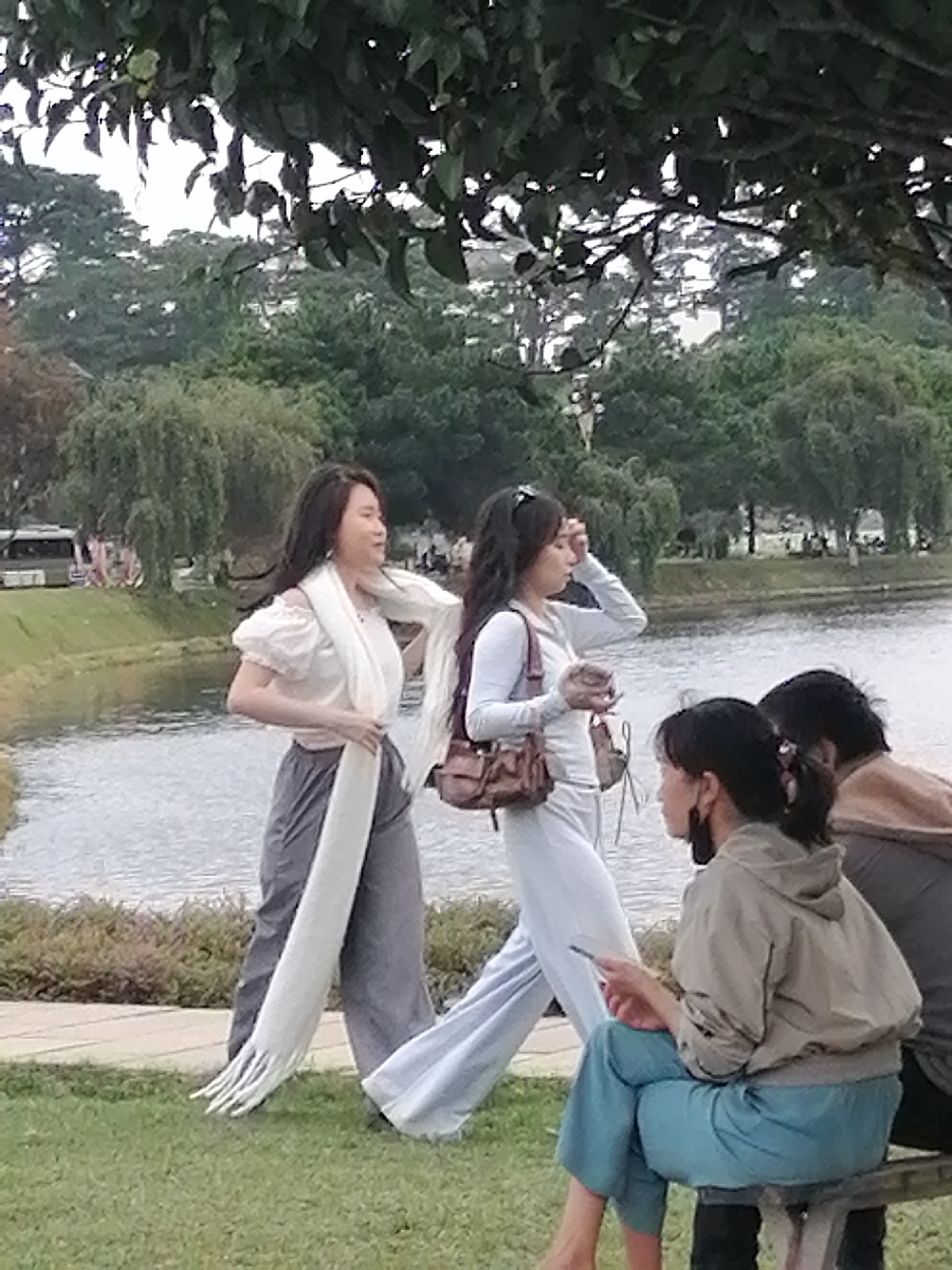 The image captures a scene at Xuan Huong Lake in Da Lat, Vietnam. In the foreground, two young women are walking, and two individuals are seated on a bench. The woman on the left is wearing a white off-the-shoulder top with puffed sleeves and gray wide-leg pants. A long white scarf is draped around her neck and trails down her front. The woman on the right is wearing a light blue long-sleeved top and matching light blue pants. She is carrying a small brown handbag. A third woman, wearing a beige hooded jacket and blue pants, is seated on a bench, looking at her phone. Another person, partially visible, is seated next to her. The background features the calm surface of Xuan Huong Lake, with trees and greenery on the opposite bank. A bus is visible on the road along the lake. The overall atmosphere suggests a casual outing in a park setting.