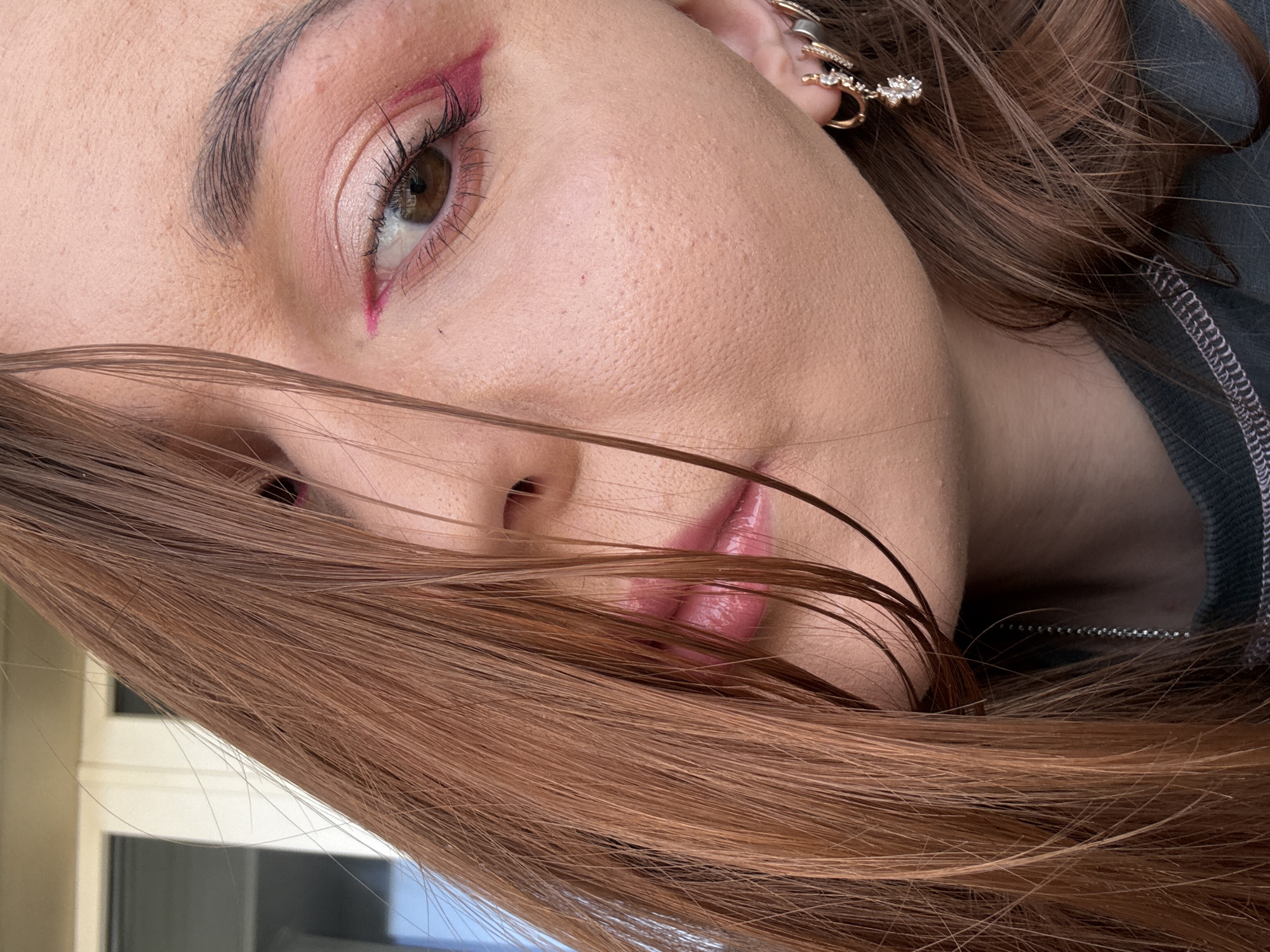 A close-up shot depicts a woman's face angled to the left, with brown hair partially covering the lower half. The woman has fair skin and brown eyes with pink eyeliner extending outward and downward from the outer corner of her eye. Her eyebrows are dark and well-defined. Her lips are visible through the hair and are glossy with a pink tint. She has multiple gold-colored earrings in her left ear. The background is indistinct, showing hints of a window and a dark gray top. There is no information to confirm that the photo was taken in Izhevsk, Russia.