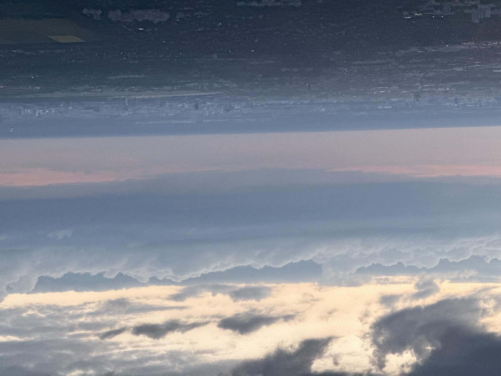 The image appears to be an inverted view from above, likely taken from an aircraft, depicting an evening or night scene. The top portion shows a darkened landscape with sparse lighting, indicative of a city or urban area at night. The presence of darker patches suggests fields or less densely populated zones. A distinct line separates this urban area from a layer of atmospheric haze or smog below, which transitions into a pinkish-purple hue. Below the haze, various layers of clouds are visible, ranging from dark gray to lighter, white-tinged formations, suggesting a low-lying cloud cover during sunset or twilight. The city is Schönefeld, Germany.