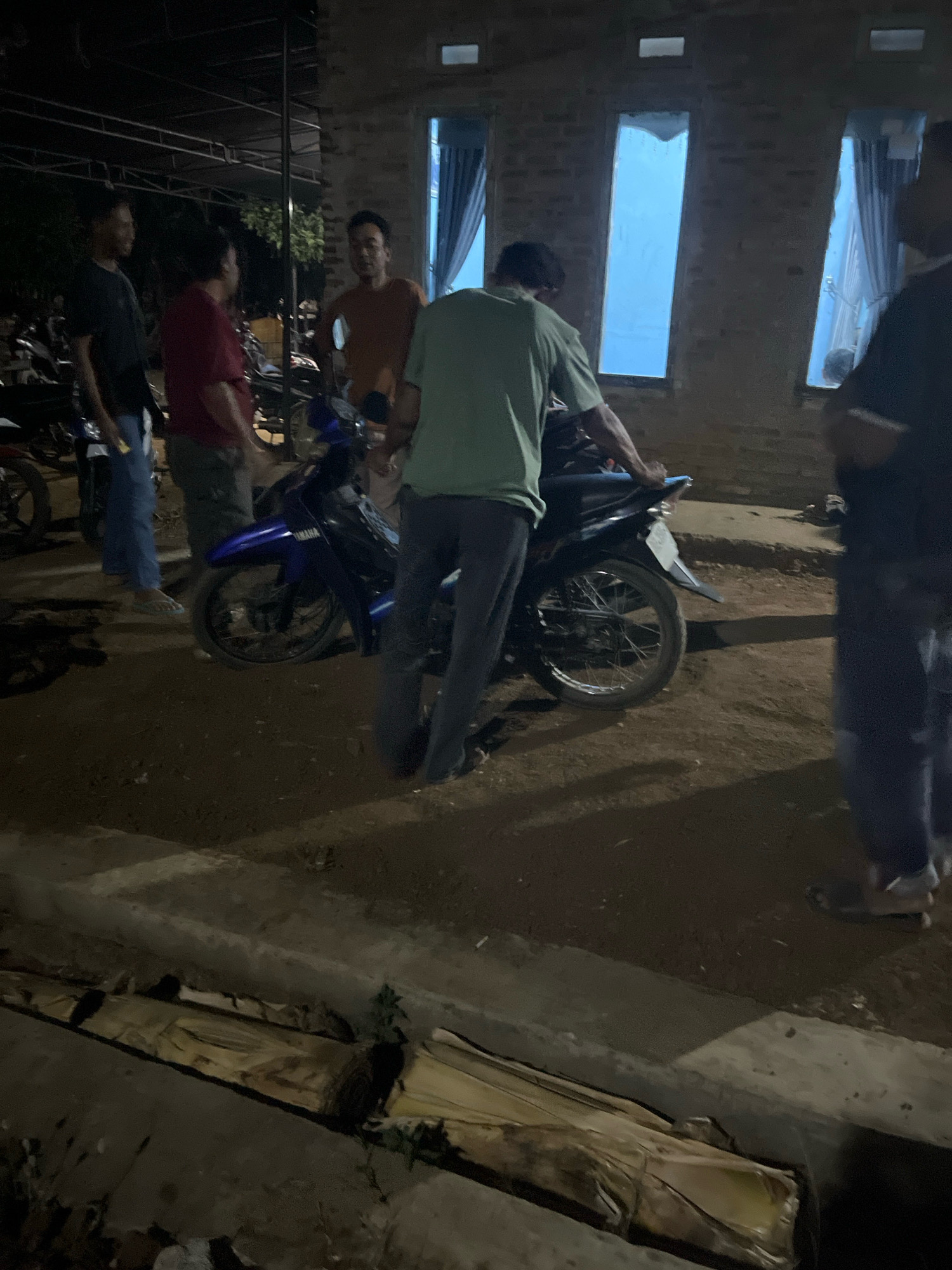 In Kebumen, Indonesia, this nighttime scene captures an outdoor gathering of at least five individuals near a brick building with open windows draped with blue curtains. A dark-colored motorcycle, possibly a Yamaha, is parked centrally. One person, wearing a green t-shirt, is seen leaning on the motorcycle, while others stand around. The ground is unpaved, with some debris and vegetation visible in the foreground. The overall lighting suggests an informal event or meeting.