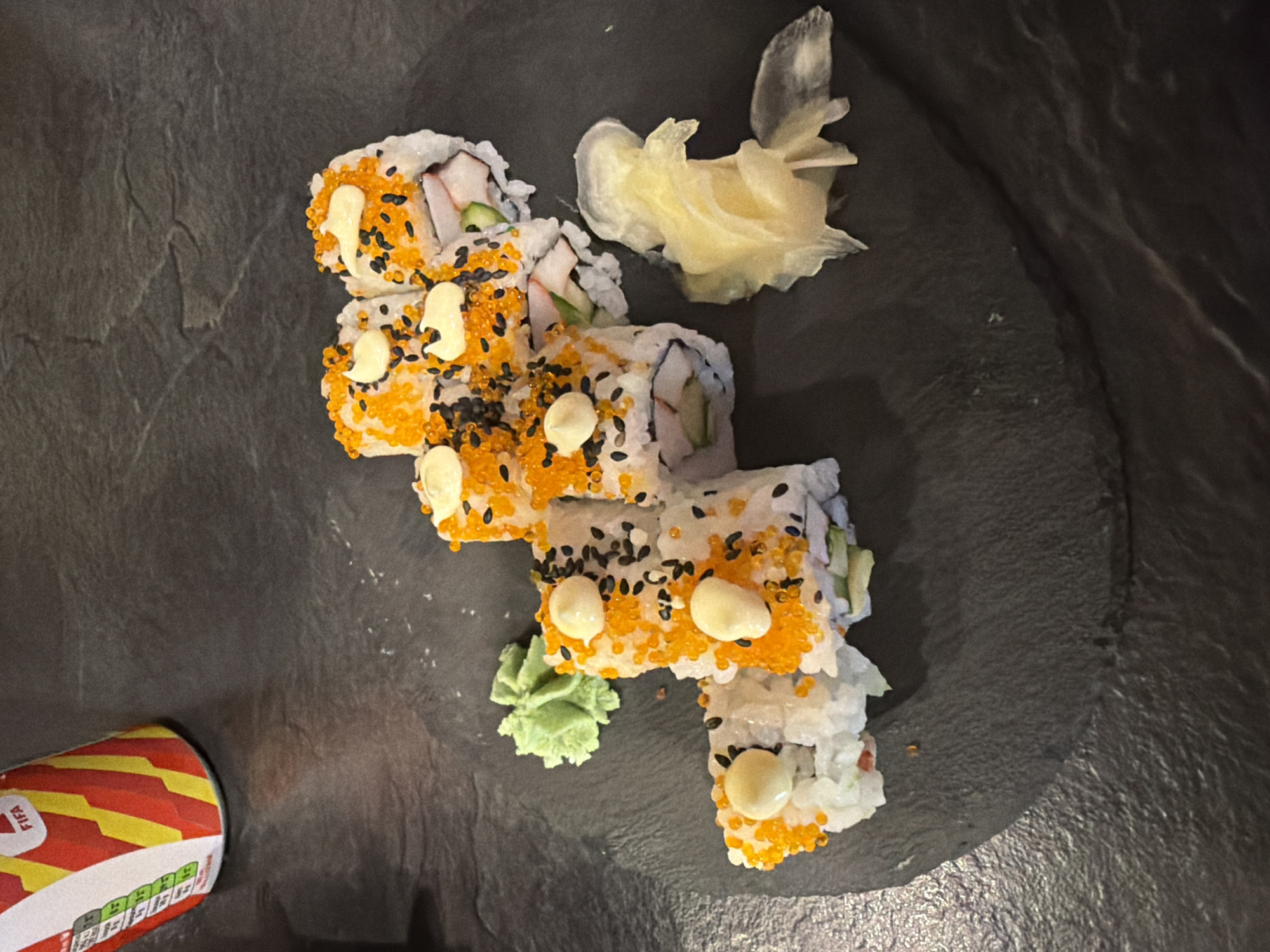 A close-up, overhead shot shows a serving of sushi rolls on a dark, textured surface, possibly a slate plate. The sushi rolls are topped with bright orange roe, black sesame seeds, and dollops of white sauce, likely mayonnaise. To the left of the sushi is a small mound of bright green wasabi. To the right, there is a pile of pickled ginger. In the upper left corner, a portion of a red, yellow, and white striped can, with "FIFA" text visible, is present. The lighting is somewhat dim, casting subtle shadows. The image does not provide any information about the city of Kiti, Cyprus, or any individuals present.