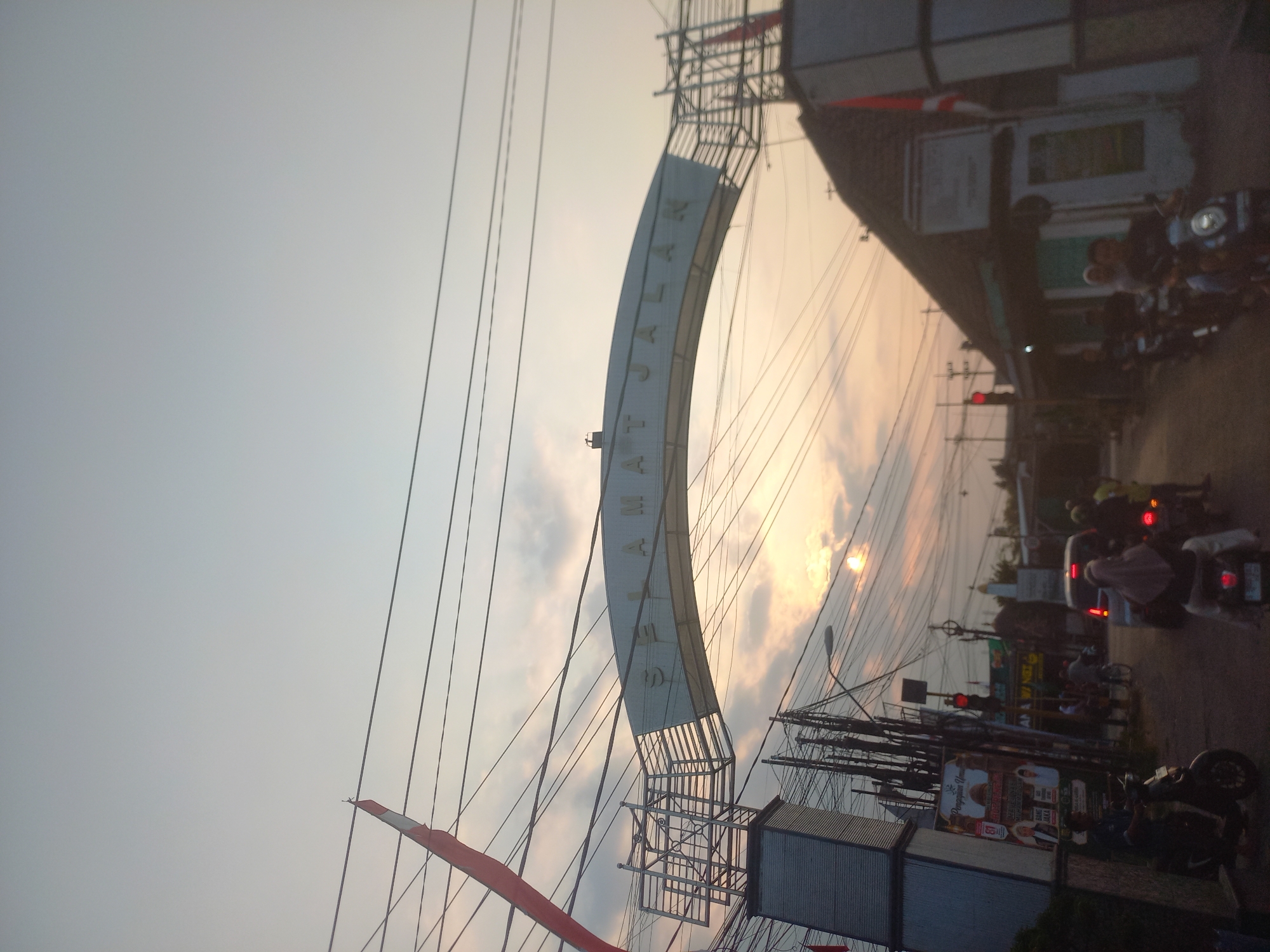 The image captures a street scene in Bedah Lawak, Indonesia, during what appears to be sunset or dawn. An arched gateway with the text "SELAMAT JALAN" (meaning "Safe Journey" in Indonesian) is prominently displayed overhead. Numerous power lines crisscross the sky above the gateway and the street. The environment suggests a busy urban or semi-urban area, with a road in the foreground and buildings lining the sides. Several motorcycles and a car are visible on the road, indicating traffic. People are riding the motorcycles, some with passengers. On the left side of the street, a large advertisement poster is affixed to a pillar supporting the gateway. A traffic light is also visible, showing red. The sky is a pale gradient of blue and orange, with some clouds, and the sun is partially visible as a bright spot peeking through the clouds. A red and white Indonesian flag is attached to a pole near the gateway on the right side of the image. The overall impression is a typical street view in an Indonesian town at a transitional time of day, with a welcoming archway.