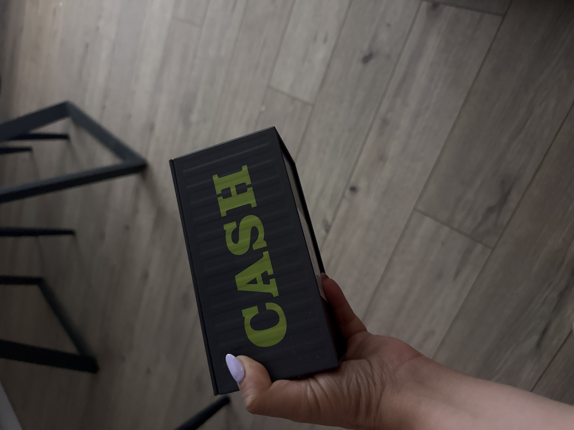 A person's hand is prominently featured in the foreground, holding a dark, rectangular box. The hand has light skin and the fingernails are adorned with a pale purple nail polish. The box itself is dark gray or black with a subtle horizontal ribbed texture. Vertically printed on the side of the box, in a vibrant lime green color, is the word "CASH."
The background reveals a light-colored floor, consistent with wood or laminate planks, laid out horizontally. In the bottom left corner, part of a dark, angular metal structure, possibly a furniture leg or frame, is visible. The lighting appears to be even and diffused, suggesting an indoor setting. No specific time of day, weather conditions, or emotional states are discernible.