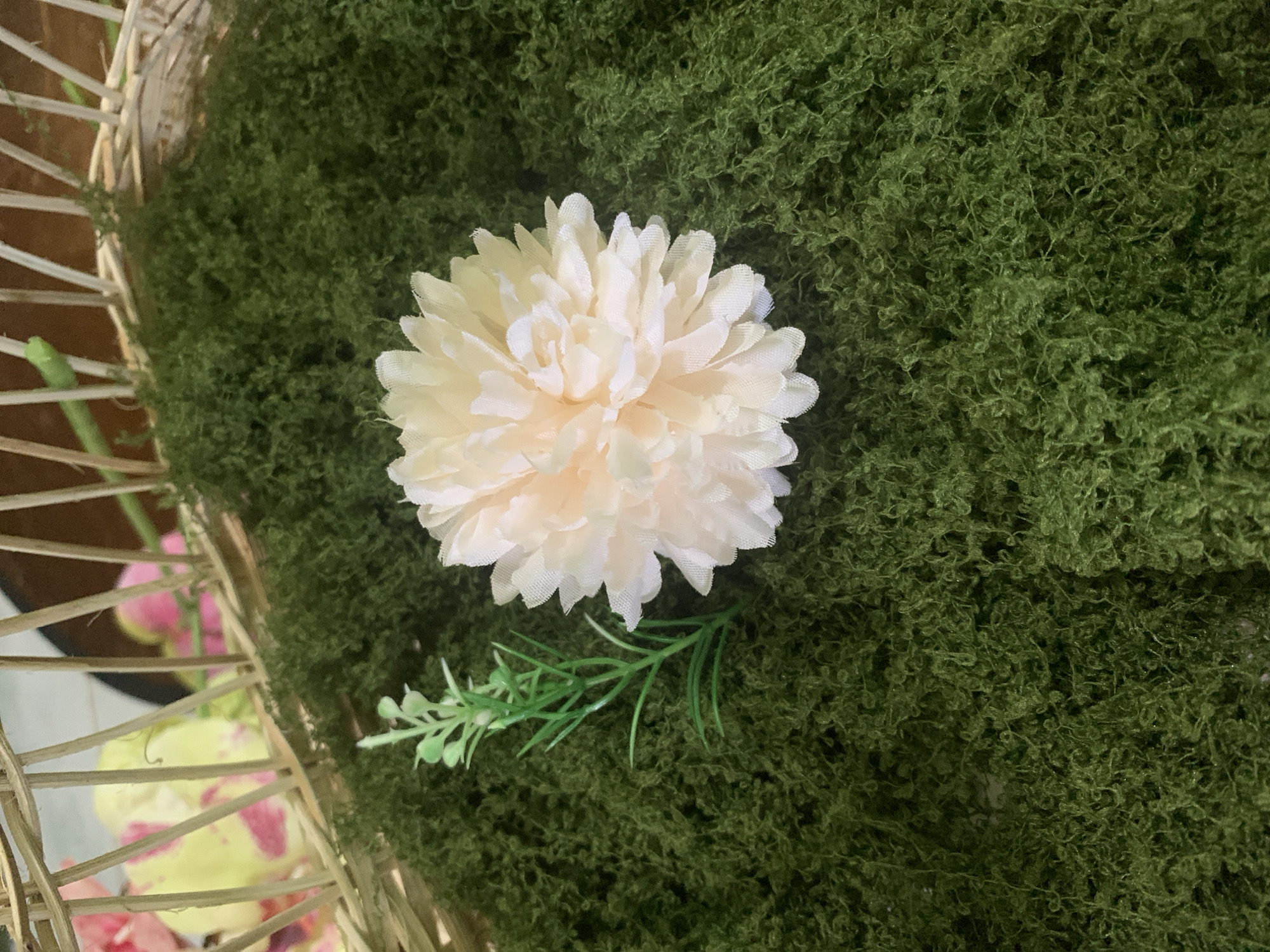 The image shows a close-up of a decorative arrangement. A light yellow, fluffy artificial flower is positioned on a bed of green, textured material that resembles moss. To the left of the flower, a small, artificial green sprig with tiny leaves is visible. In the background, parts of a woven basket are seen, with blurred pink and yellow orchids visible through its structure. The overall scene suggests a decorative display, possibly for an event or as part of a floral arrangement. There is no indication of a specific location within Kuala Pembuang, Indonesia.
