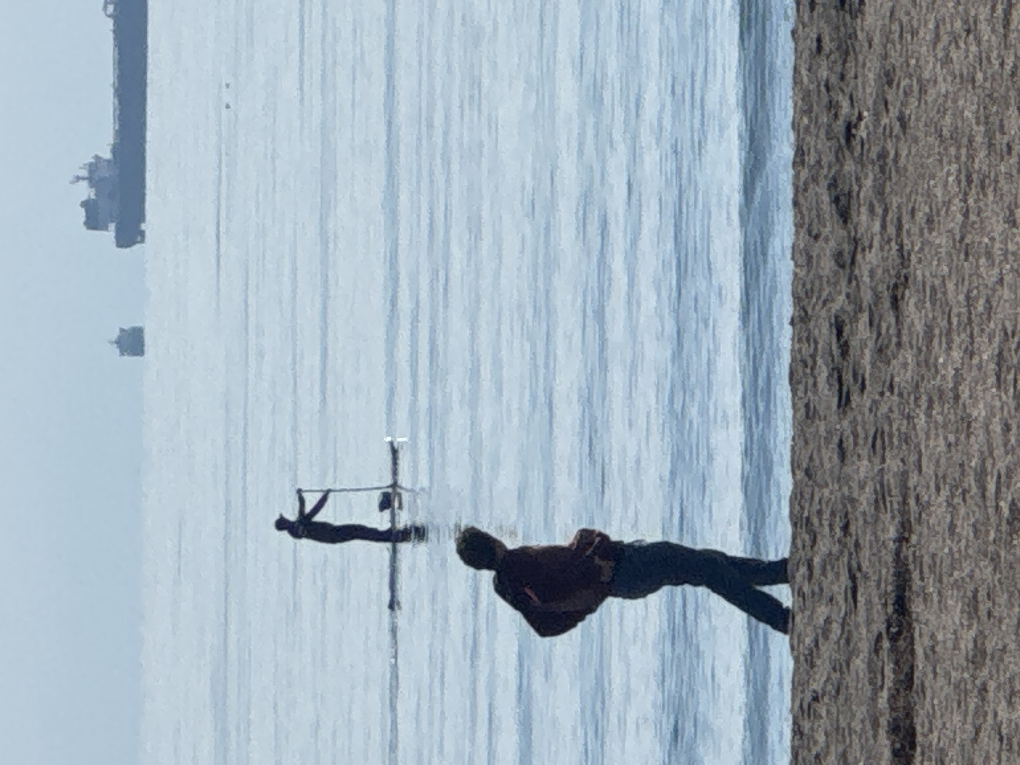 The image shows a man on a slackline over water, presumably the Black Sea, in Burgas, Bulgaria. He is wearing dark pants and a darker short-sleeved shirt. A second person, likely an aerialist, is hanging from a metal structure further out in the water. In the background, a ship and other structures are visible on the horizon. To the right of the frame is a rough textured vertical element, which looks to be from a wall or pier.