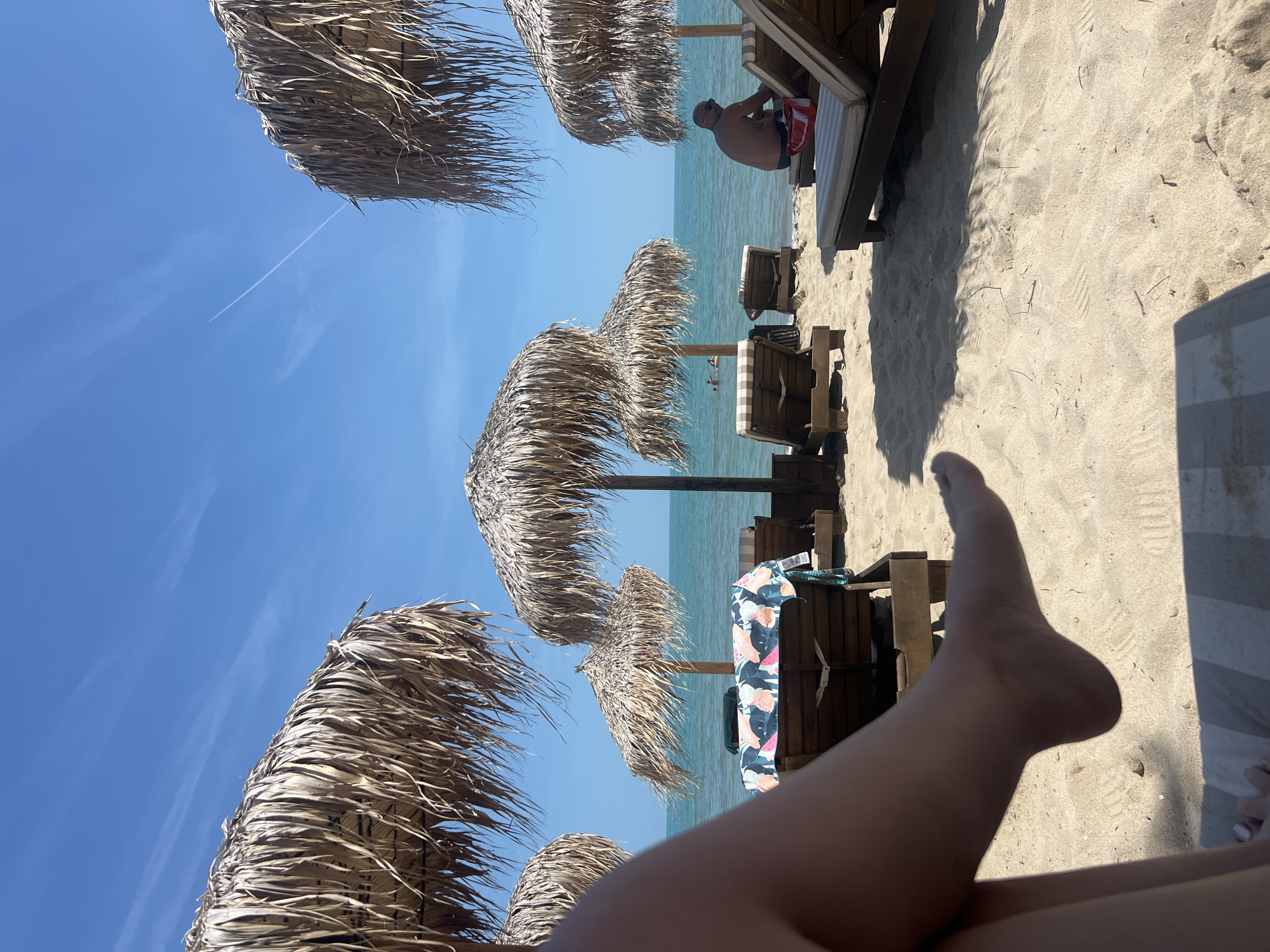 The image depicts a bright, sunny beach scene in Kranevo, Bulgaria. In the immediate foreground, the lower legs and feet of an individual are visible, positioned as if resting on a striped sun lounger, indicating a first-person perspective. The sandy beach extends into the mid-ground, where multiple wooden sun loungers with striped cushions are present. Several large, thatched straw umbrellas provide shade across the scene. One sun lounger to the left features a colorful towel with a pattern of tropical leaves and fruit. To the right, a shirtless male wearing sunglasses is seated on a sun lounger near the water's edge, facing the tranquil, blue-green sea. A red and white bag rests on the sand beside his lounger. In the background, the sea stretches to the horizon, and faint figures are discernible in the water, suggesting swimming. The sky above is clear and bright blue, featuring a single, thin white contrail from an aircraft in the upper right quadrant. The scene is illuminated by direct sunlight, creating defined shadows on the sand.