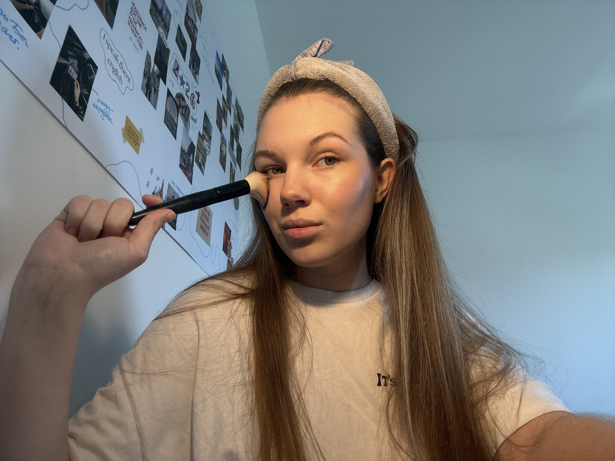 A woman is applying makeup with a brush. She is wearing a headband and a white t-shirt with the word "IT'S" printed on it. Behind her is a wall with pictures and writing on it. The writing includes the phrases "I speak fluent English" and "НЕИДЕАЛЬНОЕ ДЕЗИСТЕМЕ ИДЕАЛЬНОГО БЕЗДЕЙСТВИЯ а то!". Another written phrase reads "если бы не". The location is Vozdovac Urban Municipality, Serbia.