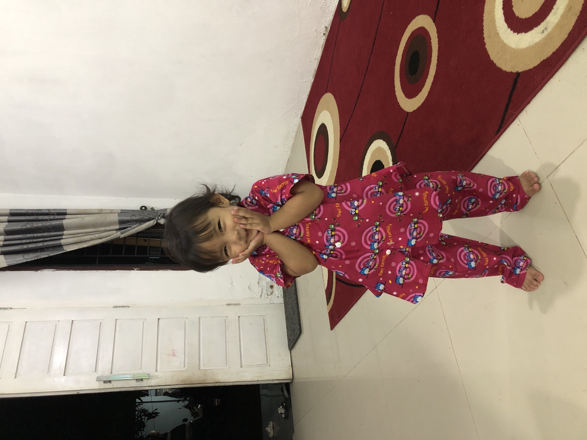 A young Indonesian girl stands in a doorway, her hands cupped to her cheeks in a playful pose. She wears pink pajamas adorned with a repeating owl family pattern. The girl is positioned on a tiled floor, with a red rug featuring concentric circles to her right. Behind her, a white door with paneling stands ajar, revealing a glimpse of another room. To her left, a dark opening suggests an exterior view, possibly a street in Pashiebeutong, Indonesia. A striped curtain hangs near the doorway. The lighting appears to be from an interior source, illuminating the scene.