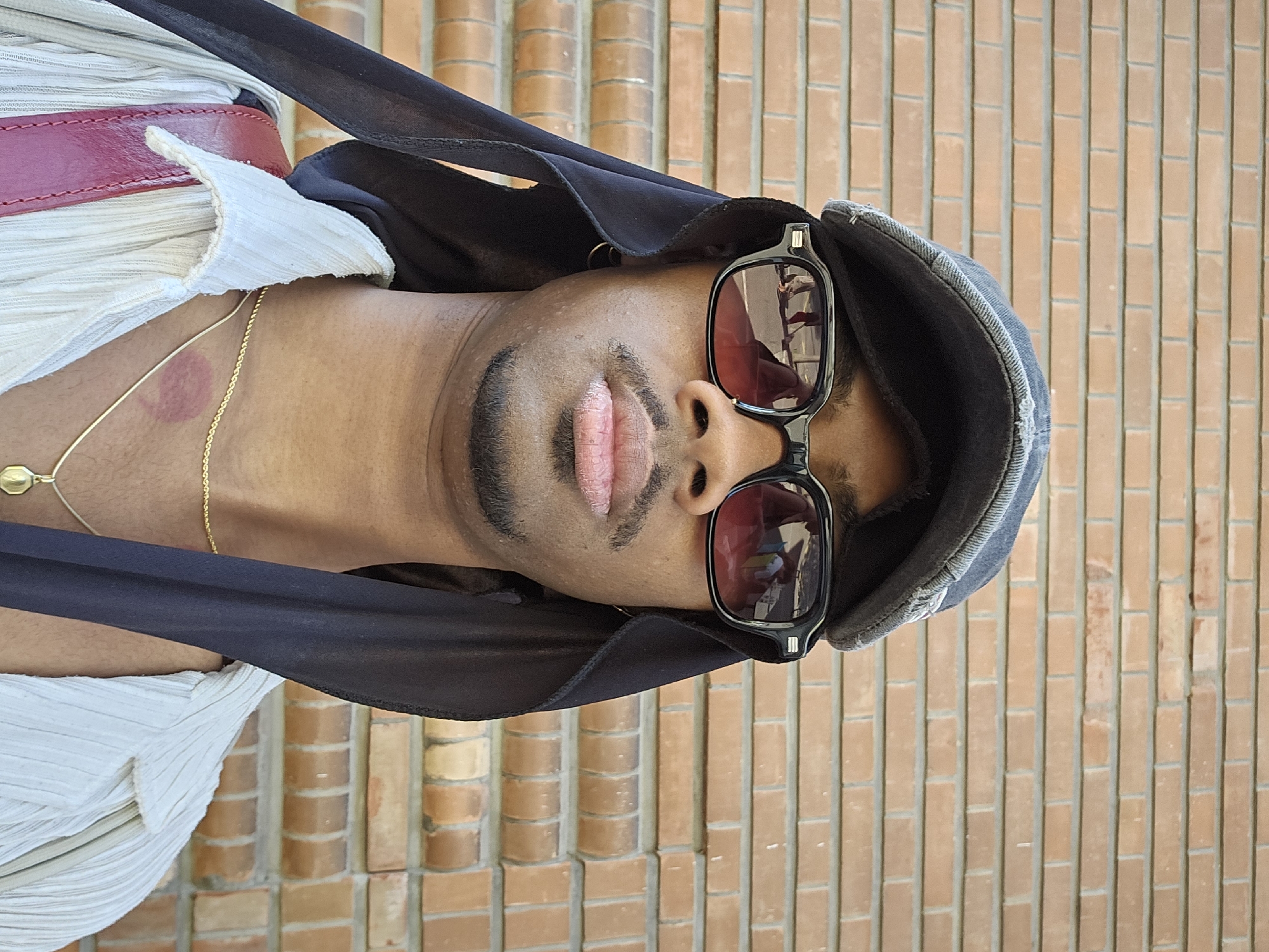 A man is photographed in front of a brick wall. He is wearing sunglasses, a headscarf, and a light-colored shirt with a red belt. There is a lipstick mark on his neck. The photo was taken vertically and is rotated. No location in Massy, France can be inferred from this image.