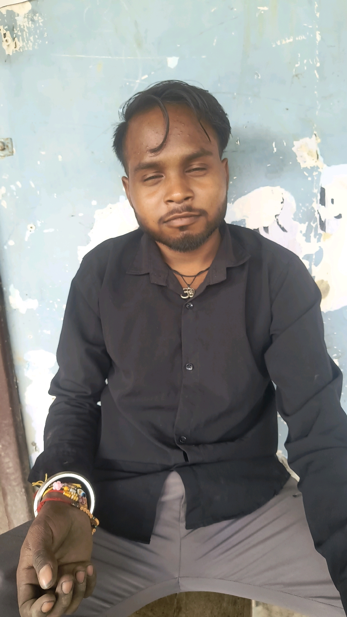 A person with dark skin and black hair is sitting with their eyes closed, wearing a black collared shirt and gray pants. A black string necklace with a pendant is visible on their chest. The person's left hand rests on their lap, adorned with several bracelets. The background is a textured, light blue wall with patches of peeling paint. The scene appears to be an outdoor setting, possibly in Loni, India.