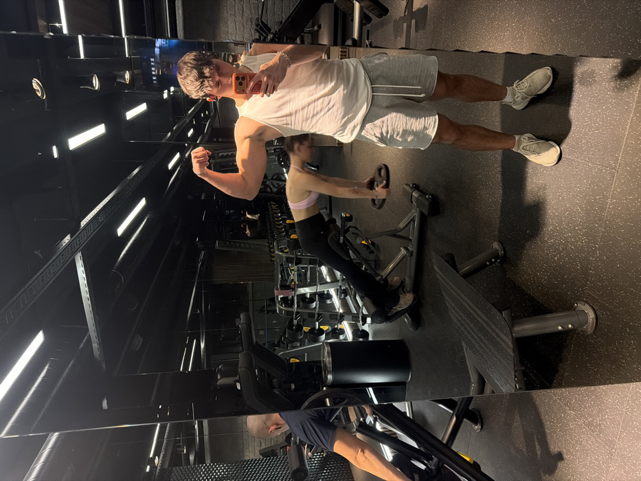 An interior view of Debute Fitness, located in Sofia, Bulgaria, depicts a male individual taking a mirror selfie. The male subject, positioned in the foreground, wears a white tank top, grey athletic shorts, and light-colored athletic shoes. He is flexing his right bicep while holding a coral-colored smartphone in his left hand to capture his reflection. A small earpiece is visible in his right ear, and a silver-colored bracelet on his left wrist.
The gym features a dark aesthetic with a speckled rubber floor, dark reflective wall panels, and an exposed black ceiling with visible ductwork and horizontal fluorescent light fixtures.
In the background reflection, a female individual is performing an exercise on a hyperextension machine. She is wearing a light pink sports bra and black leggings, holding small black weight plates near the floor. Further to the left in the reflection, another male individual, bald and wearing a dark blue t-shirt, is seated at a leg press machine. Various gym equipment, including weight racks and a black cylindrical trash bin, are visible throughout the space. There are no direct interactions observed between the individuals.
