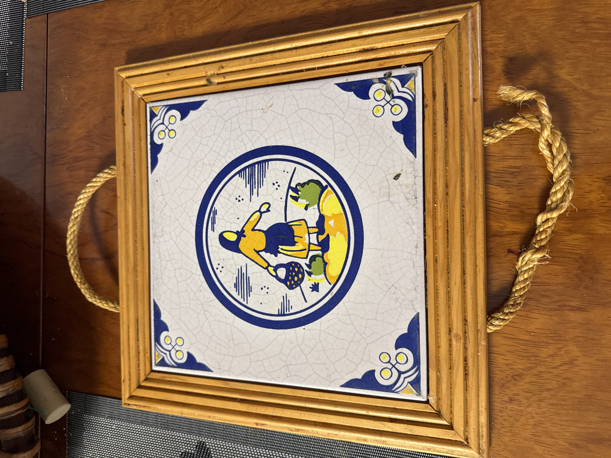 The image shows a square ceramic tile framed in wood, hanging against a wooden surface. The tile depicts a circular scene with a stylized woman in a yellow top and blue skirt carrying a basket, standing on a green and yellow ground with blue accents. The tile is set in a light-colored wooden frame with a ribbed texture. A thick, braided rope is attached to the top of the frame, suggesting it is a hanging decoration. The wooden surface below the tile is a rich, polished brown. The overall impression is that of a decorative item displayed indoors. There is no indication of the city of Gandia, Spain, or any other specific location.