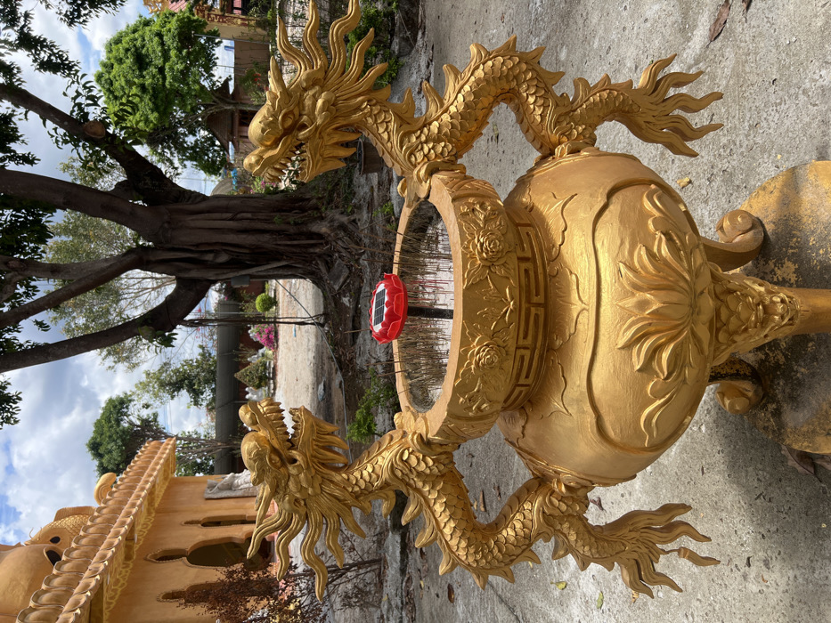In the city of Đăk Mar, Vietnam, a large, ornate golden incense burner stands in the foreground of an outdoor scene. The burner is fashioned with two elaborate golden dragon statues acting as handles, their heads facing outward from the central basin. The basin contains numerous thin, dark, vertically oriented incense sticks embedded in a substrate, with a red, lotus-shaped solar-powered light fixture positioned centrally among them. The burner's body features sculpted lotus petals and rests on three legs, each terminating in a dragon head motif. The immediate ground is concrete. To the left, a multi-tiered ochre-colored temple building with intricate architectural details and several white statues is partially visible. In the midground, a large, gnarled tree with exposed roots dominates the center-right, beyond which lies an open area with trimmed bushes, some displaying pink flowers, and additional white statues. The background features a partially cloudy sky and dense foliage. No individuals are present in the scene.