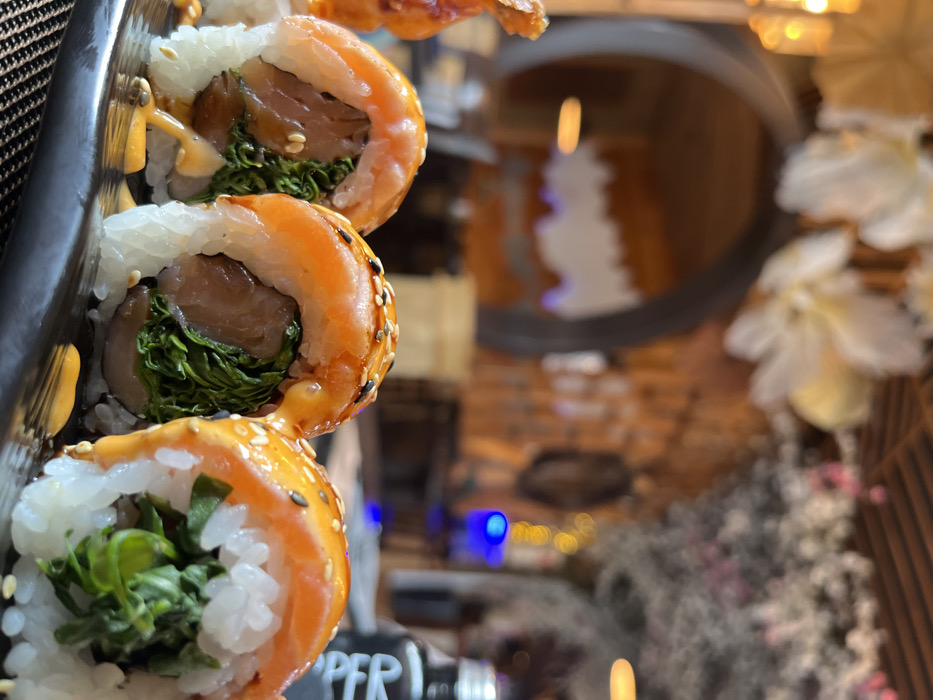 The image presents a close-up, shallow-depth-of-field view of three sushi rolls arranged on a dark, rectangular serving plate, positioned on a dark, woven placemat. Each roll is composed of white rice, filled with green leafy vegetables and a piece of brown fish, topped with a slice of raw salmon, drizzled with an orange-yellow sauce, and sprinkled with white and black sesame seeds. Additional sauce is visible on the plate.
The background, significantly blurred, depicts a restaurant interior. A large, dark, arched entryway occupies the mid-left, revealing a warm-toned room with a distinct, jagged white decorative element against a darker wall. To the right of the arch, a wall features vertical wooden slat paneling. Blurred white and pink floral decorations are suspended throughout the ceiling area. Multiple diffused yellow light sources and a prominent blue light are visible as bokeh effects. On the far right, a dark bottle with partial white lettering, "PPER," is present on a surface.
This scene is located within the Tokyo Grill & Sushi restaurant in Bad Sobernheim, Germany.