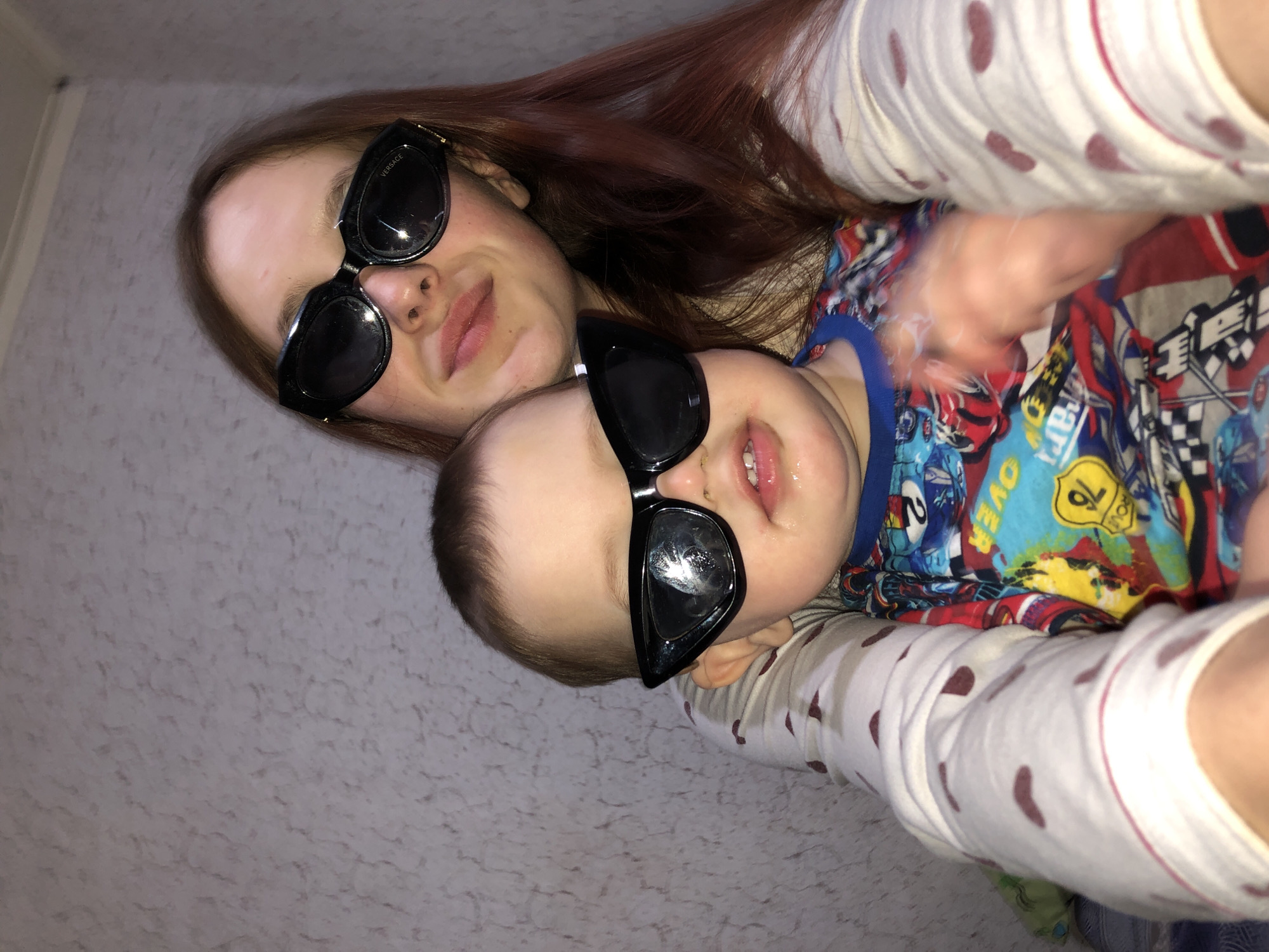 A photo of a woman and a baby, both wearing sunglasses, possibly in a domestic setting. The woman has auburn hair and the baby is wearing a colorful shirt with a blue neckline. The baby's face is slightly drooling. The woman and baby appear to be interacting. The location in the city of Фоки, Russia is not visible or implied by the available evidence.
