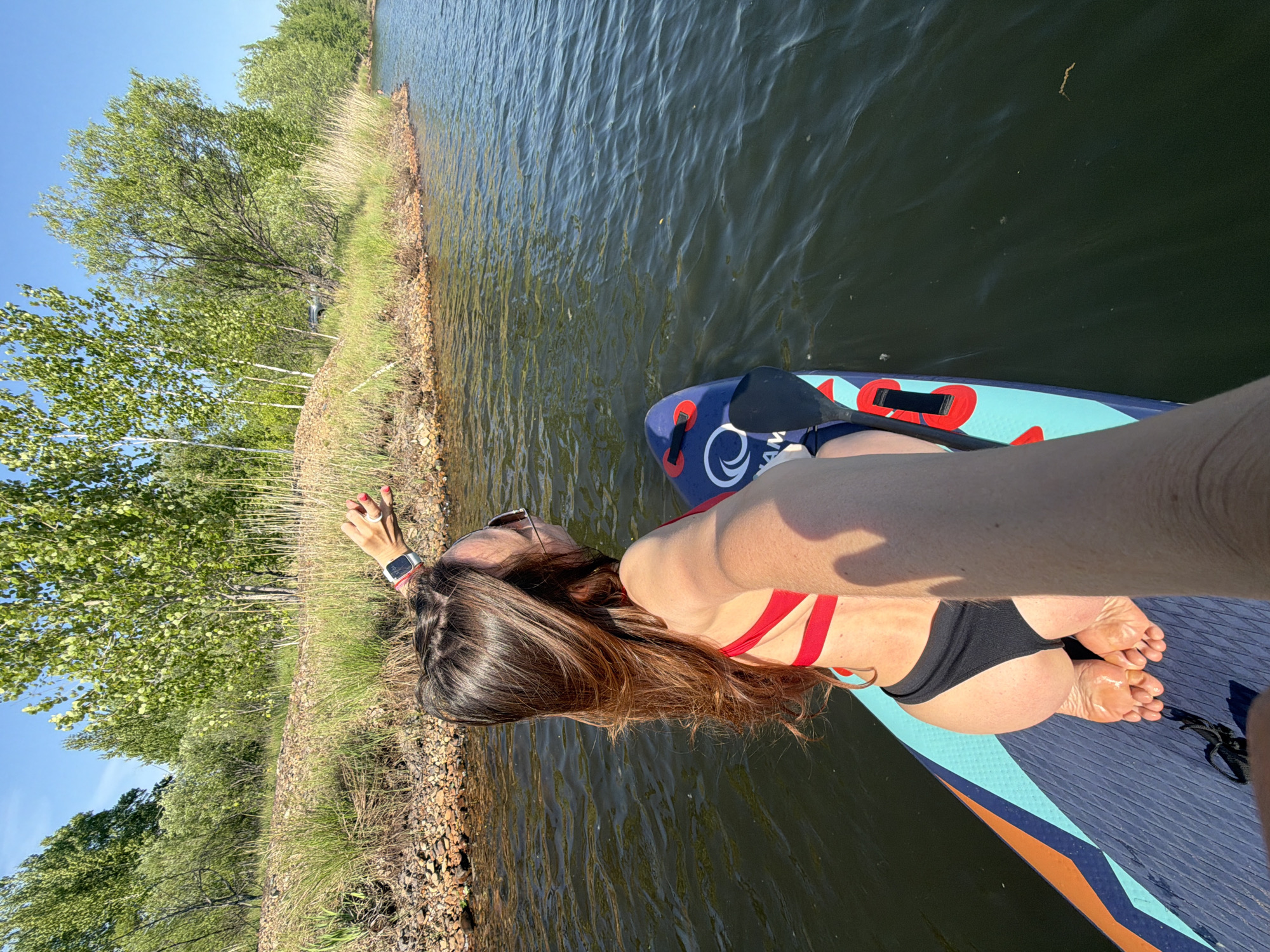 A woman in a red swimsuit top and black bottoms is lying face down on a paddleboard in a body of water. She is wearing sunglasses and a smartwatch. Her hair is long and brown. The paddleboard is blue and teal with orange accents. The water is dark and reflective. The shoreline is visible with trees and vegetation. The photo appears to be taken from the perspective of someone else on the paddleboard. The location is likely a lake or river near a wooded area, but cannot be pinpointed to Birobidzhan.