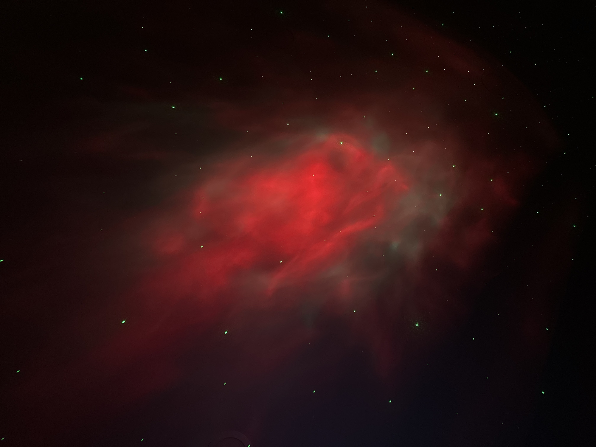 The image shows a nebula in a dark sky. The nebula is primarily red with hints of green, resembling a cloud-like structure. Small, green dots are scattered across the image, likely representing stars. The background is dark and empty.
There is no evidence in the image to support a location in the city of Бараўлянскі сельскі Савет, Belarus, or any information about people, subjects, objects, actions, or interactions.