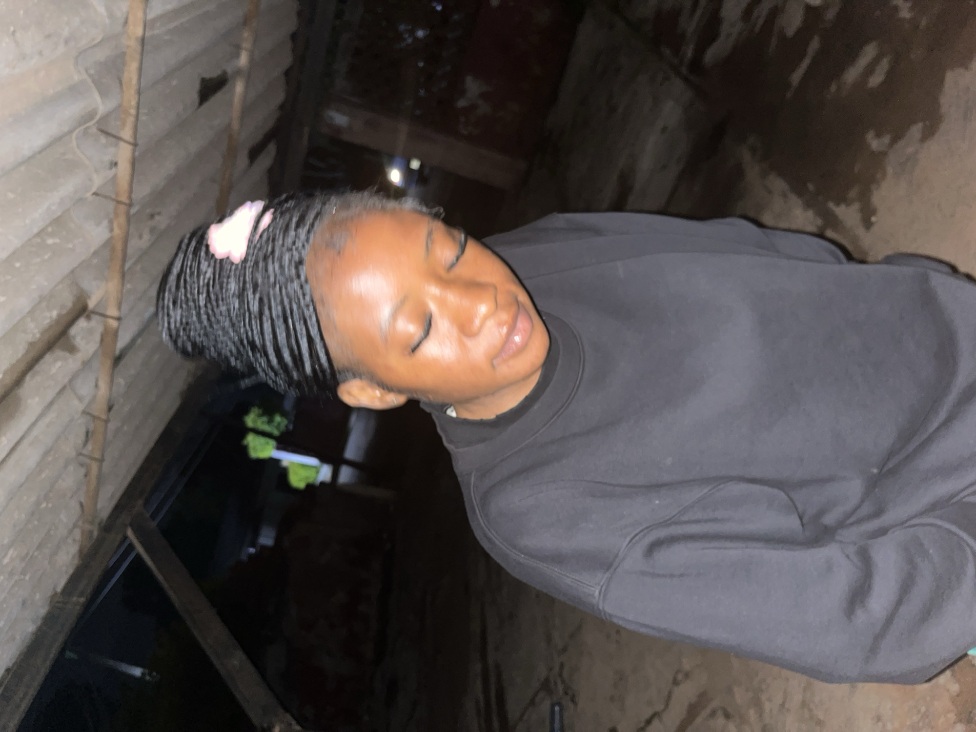 The image depicts a young Black woman with braided hair adorned with a pink accessory, standing in what appears to be an outdoor, possibly semi-enclosed, area. She is wearing a dark-colored sweatshirt. Her eyes are closed, and her head is slightly bowed, suggesting a moment of rest or contemplation. The background shows a rustic ceiling made of corrugated material supported by wooden beams, indicating a possibly unfinished or utilitarian structure. The ground appears to be dirt or sand, with some patches of moisture. The lighting suggests it might be evening or nighttime, with artificial light sources creating shadows. The overall environment seems to be in a less developed or rural part of the city of Jos, Nigeria, characterized by simple construction and natural ground cover.
