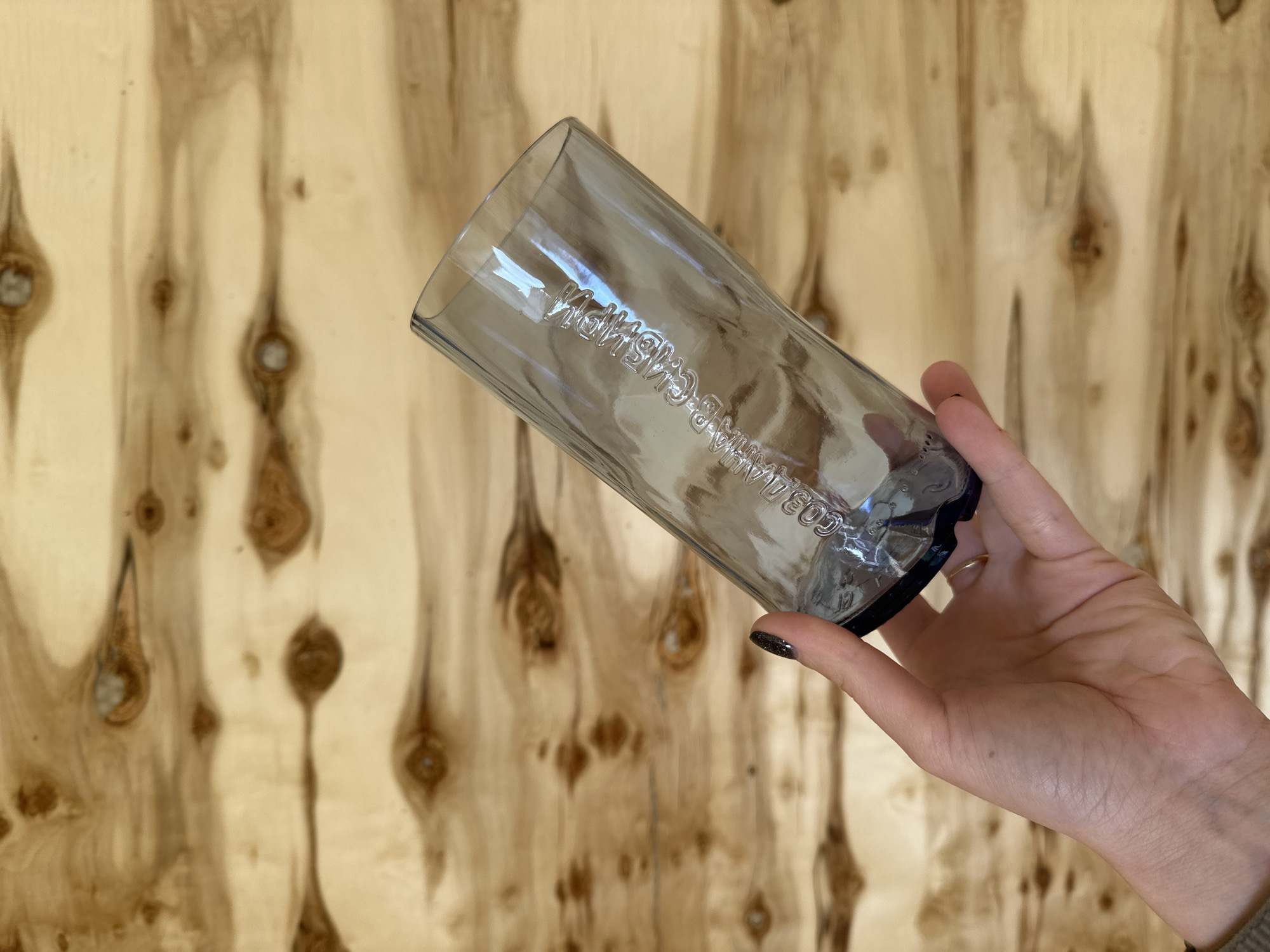 A human hand holds a transparent, smoky-tinted glass against a light wooden background. The wood features prominent vertical grain patterns and numerous dark knots, suggesting a plywood panel or wall. The hand, with dark, glittery nail polish visible on the index finger and a plain gold-colored ring on the ring finger, holds the glass tilted. Embossed or debossed text is visible on the side of the glass. Due to the glass's transparency and angle, the Cyrillic text is mirrored and slightly distorted, but it clearly reads "СОБСТВЕННОЙ ИСТОРИИ" (Sobstvennoy Istorii), which translates to "OWN HISTORY" or "PERSONAL HISTORY". The scene appears to be indoors. The location context is Kuda, Russia.