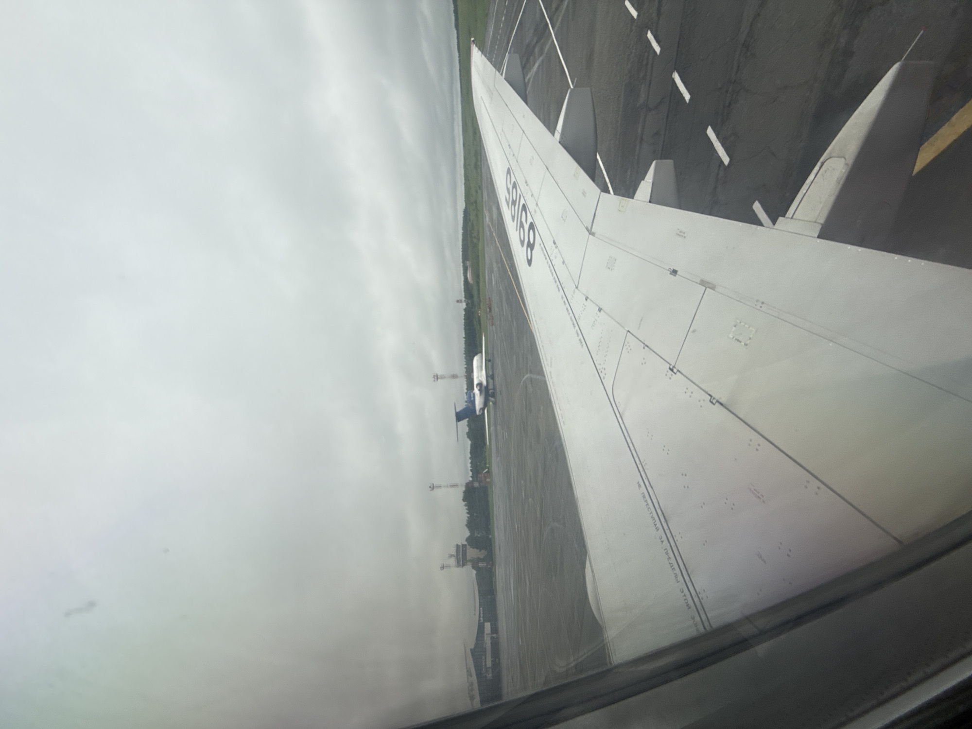 A photograph taken from the window of an airplane. The wing of the aircraft dominates the foreground on the right. Through the window, we see the airport tarmac, grass, and a cloudy sky. In the distance, there is a control tower and another plane. The image appears to have been taken at the airport in Новое Мартьяново, Russia.