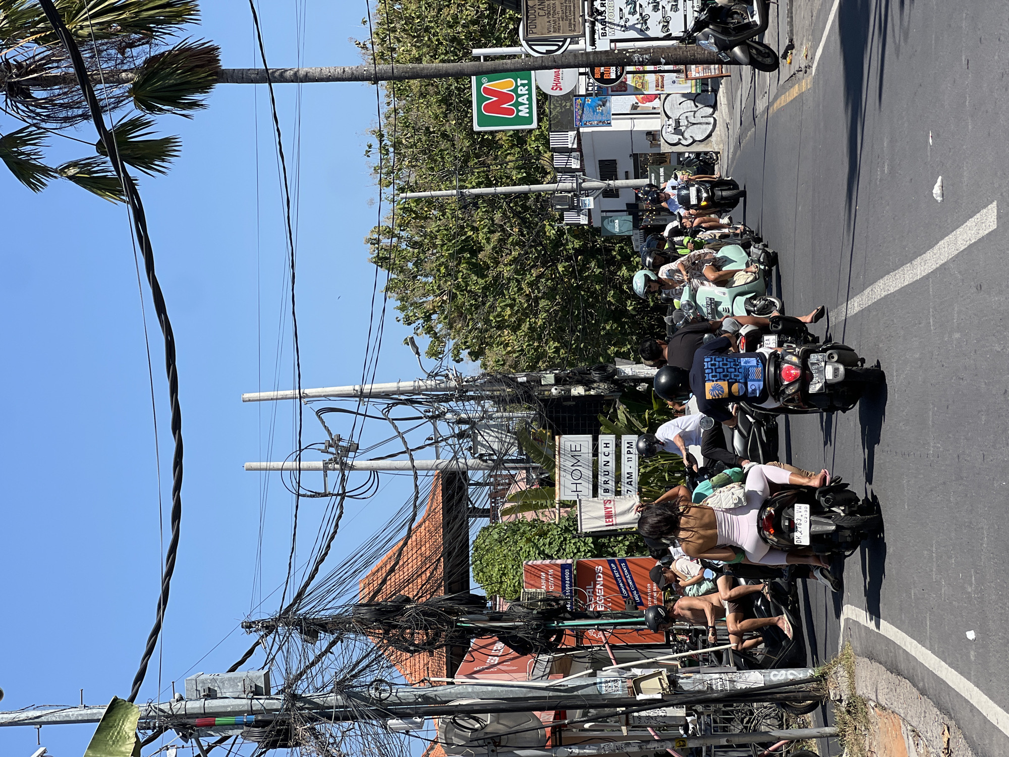 The image shows a street scene in North Kuta, Indonesia, on a sunny day with a clear blue sky. A procession of motorcycles and scooters navigates the asphalt road, lined with buildings, palm trees, and a dense network of overhead power lines. On the left side, a multi-story building with a red tile roof is visible, adorned with various signs, including one for "LEGAL LEGENDS" and another with "HOME." A green awning hangs from a utility pole. Further down the street, a prominent "M MART" sign with its distinctive red and green logo stands out. The road is marked with white and yellow lines. Several individuals are riding on the motorcycles, some wearing helmets. The overall impression is one of bustling street activity in a tropical urban environment.