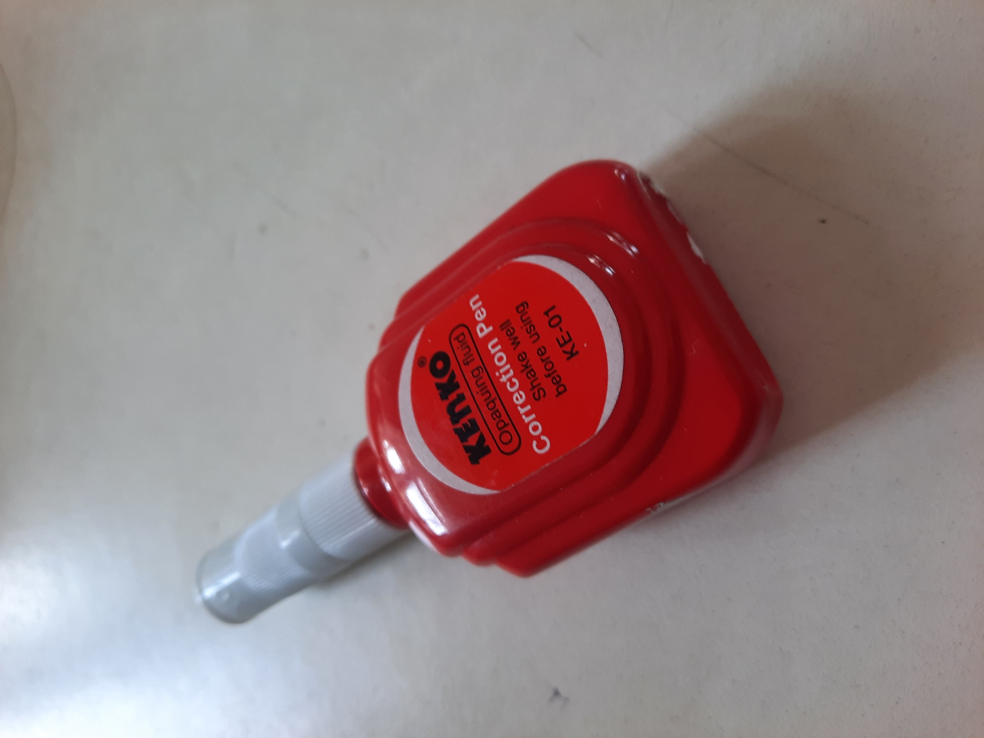 A close-up, eye-level, medium shot captures a red correction fluid pen with a white tip, resting on a light-colored, textured surface, likely a desk or tabletop. The pen is angled with the tip pointing upwards and slightly to the left. The red plastic body of the pen is glossy, reflecting the ambient light. A white label is affixed to the pen's body, featuring black text. The label reads "Kenko" at the top, followed by "Opaquing fluid" and then "Correction Pen". Below that, smaller text advises "Shake well before using", and at the bottom, "KE-01". The background is out of focus, revealing a light, neutral environment with soft shadows. The scene's lighting is even and diffused. The image lacks any discernible elements that would indicate its location in Lhokseumawe, Indonesia.