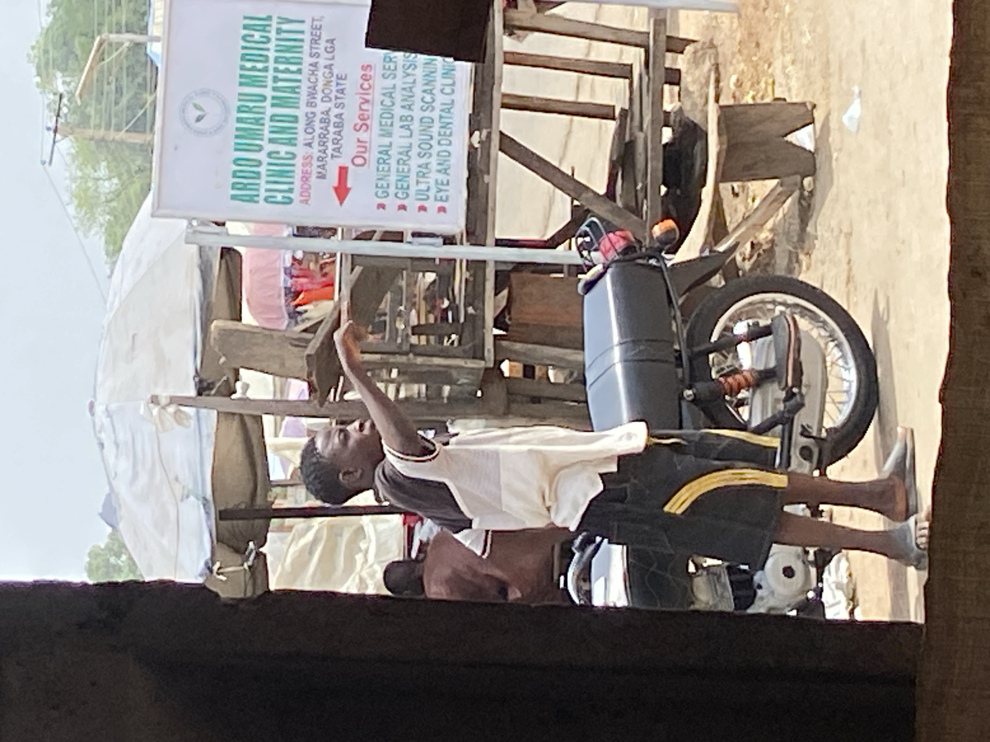 The image captures an outdoor scene in Mararaba, Nigeria, featuring a young man and a medical clinic signboard as central elements. The primary subject is a dark-skinned young man, likely a teenager or young adult, standing in the mid-ground. He wears a two-toned t-shirt (white over dark grey/black) and dark shorts with yellow stripes. His right arm is raised, possibly gesturing or pointing towards something out of frame. He wears light-colored flip-flops. Another person is partially visible behind him.
To the right of the young man is a black motorcycle, parked and visible from its front wheel and engine area. The ground appears to be unpaved, possibly dirt or gravel. In the background, various wooden structures, possibly market stalls or temporary shelters, are visible, some covered with light-colored fabric or tarpaulin.
A prominent white signboard is positioned upright, advertising "ARDO UMARU MEDICAL CLINIC AND MATERNITY." The address is clearly stated as "ALONG BWACHA STREET, MARARRABA, DONGA LGA TARABA STATE." Under the heading "Our Services," the sign lists:
- GENERAL MEDICAL SERV
- GENERAL LAB ANALYSIS
- ULTRA SOUND SCANNIN
- EYE AND DENTAL CLINIC
The scene is brightly lit, suggesting it is daytime with clear weather. The overall impression is a bustling, everyday street or market environment in Mararaba.