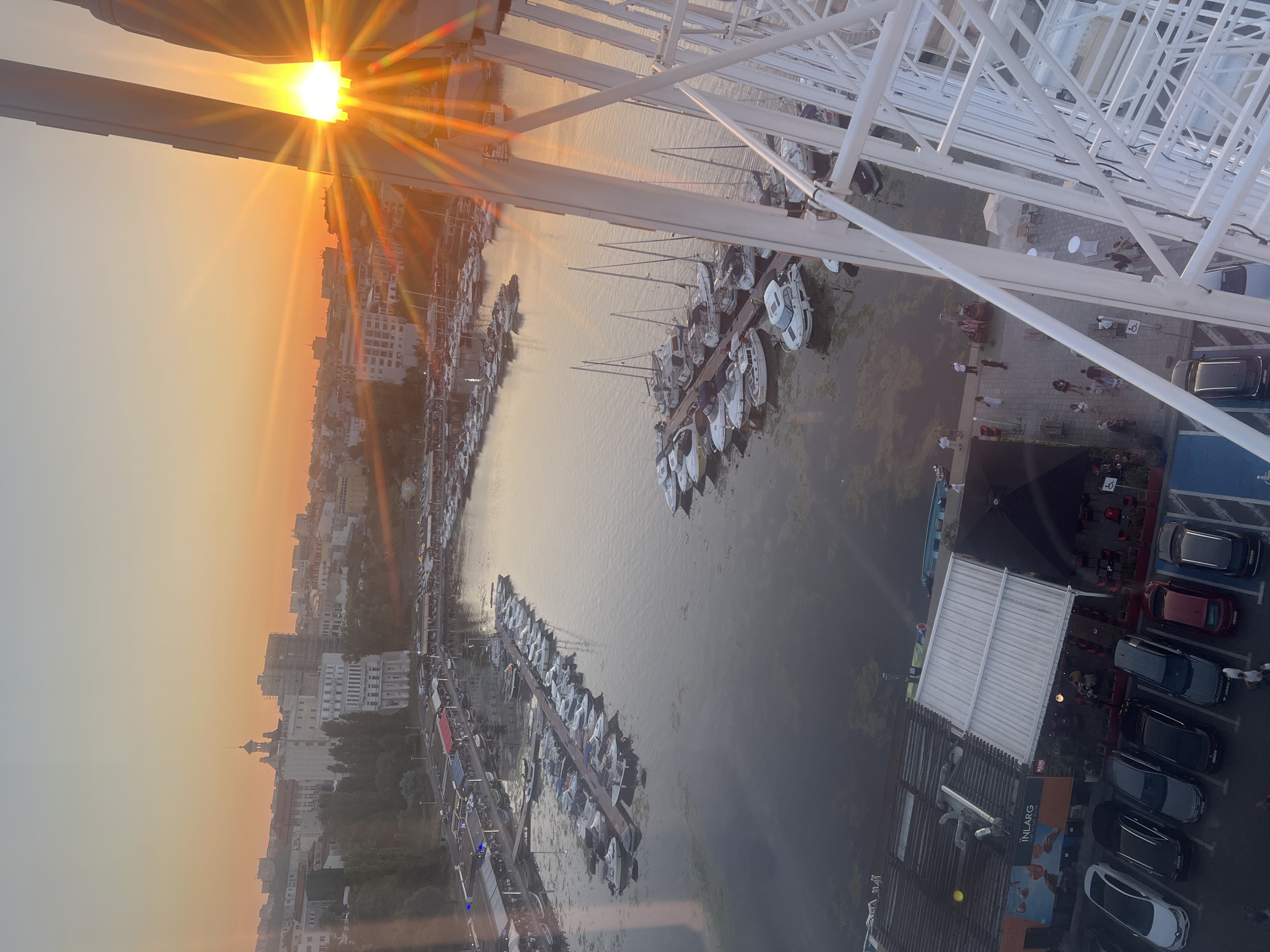 The image captures a panoramic view of a marina in Constanța, Romania, during sunset. A large Ferris wheel, white in color, dominates the right side of the frame, its structure creating geometric patterns against the sky. The setting sun, a brilliant orange orb, is partially obscured by the Ferris wheel's spokes, casting a warm glow and lens flares across the scene.
Below, the marina is filled with numerous boats of various sizes, predominantly white, docked in neat rows. The water's surface reflects the warm hues of the sunset and the surrounding buildings. Along the waterfront, there are several buildings with awnings and outdoor seating areas, suggesting restaurants or cafes. People can be seen walking along the docks and seated at tables, enjoying the evening ambiance.
In the background, the cityscape of Constanța unfolds. Buildings of different architectural styles, some modern and others older with distinct rooftops, are visible against the soft gradient of the sunset sky, which transitions from a bright orange near the horizon to a pale yellow and then a light blue higher up. Trees line the waterfront, adding a touch of greenery to the urban landscape. The overall scene is tranquil and picturesque, highlighting the beauty of the coastal city at dusk.