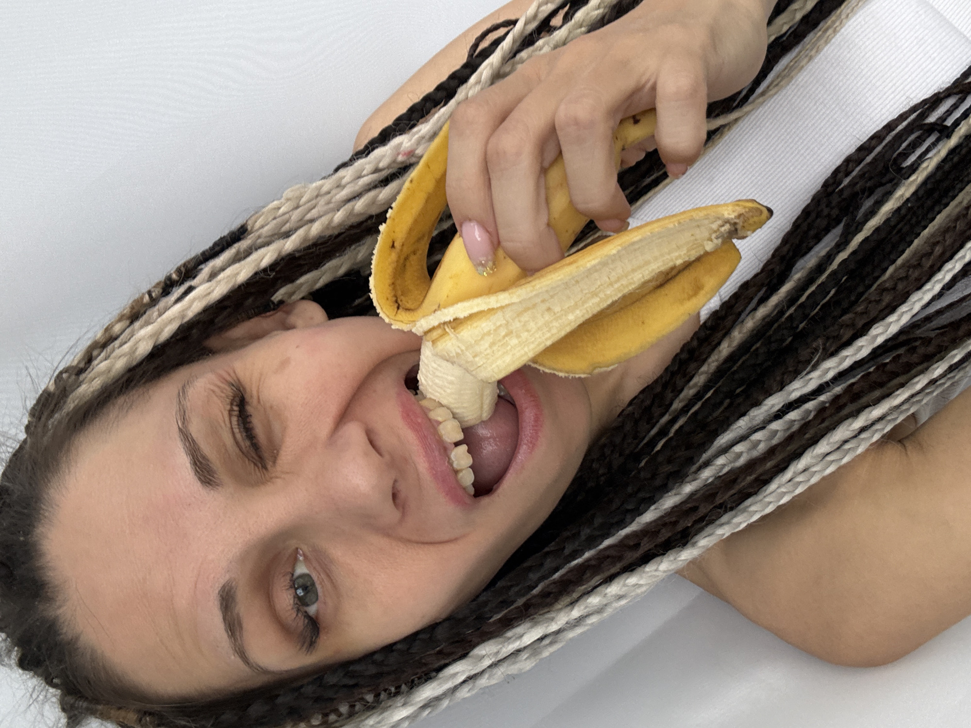 The image captures a close-up, angled shot of a woman interacting playfully with a banana. Her head is tilted, giving the impression she is lying down, and she is looking directly at the viewer with a wide, toothy smile. Her right eye is partially closed, suggesting a wink or an expression of amusement.
Her hair is a prominent feature, styled in numerous long, thin braids or dreadlocks that vary in color, including dark brown/black, medium brown, and light/blonde. One hand is visible, holding the yellow, partially peeled banana. This hand has light pink nail polish with a glitter accent on the nails. The banana's fruit is positioned inside her open mouth, as if she is about to take a bite or is playfully posing.
The background is a plain, bright white surface, suggesting an indoor setting with clean, even lighting. There are no other objects, people, or visible text in the frame. This scene takes place in Rusanovo, Russia.