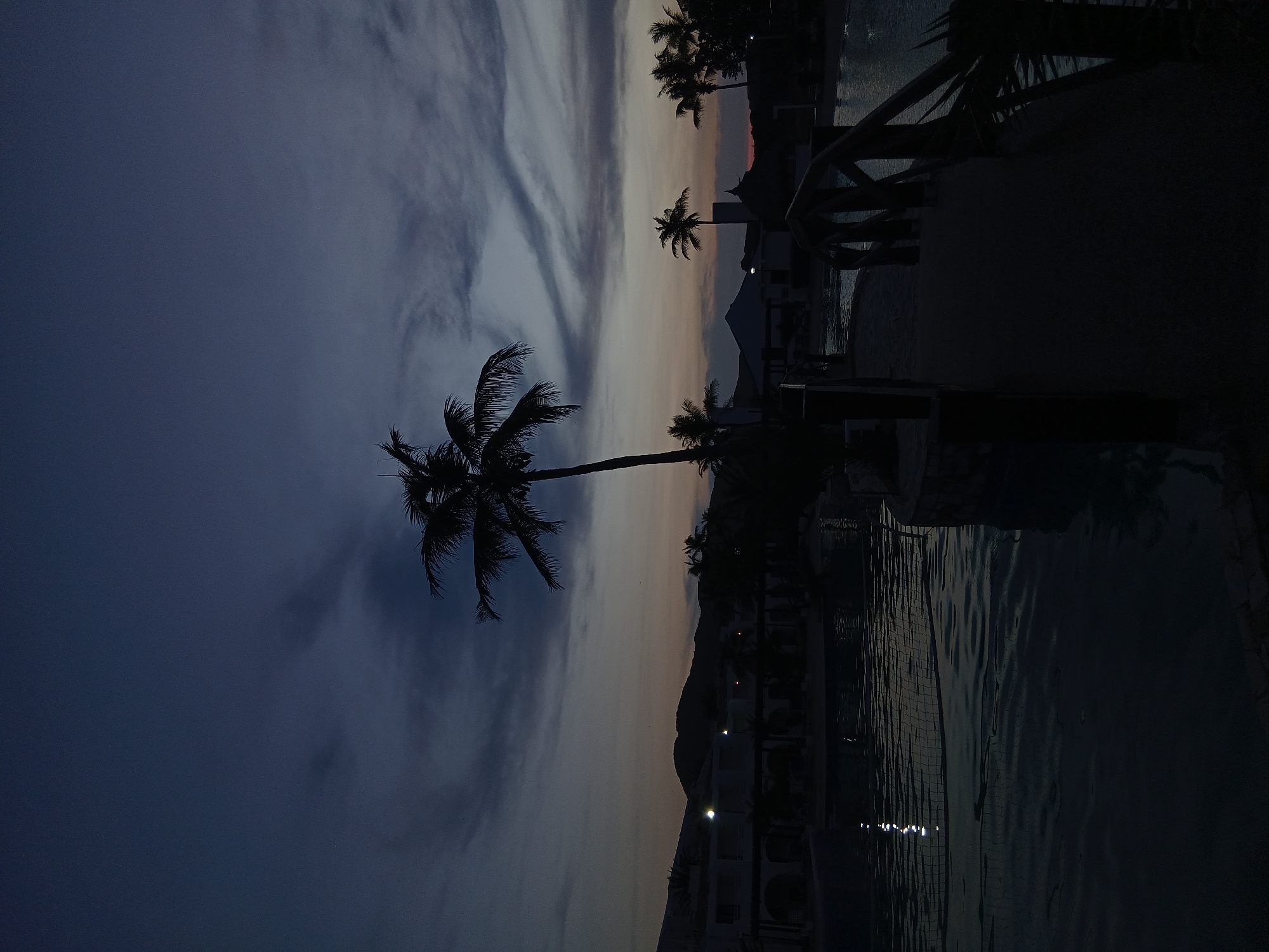 The image depicts a dimly lit scene in Altagracia, Venezuela. The sky exhibits a transition from dark blue to lighter shades, suggestive of dusk or dawn, with scattered clouds. Palm trees are silhouetted against the sky. Adjacent to the trees, a body of water, likely a pool, reflects the ambient light. Buildings are visible in the background, and a silhouette of mountains rises in the distance.
