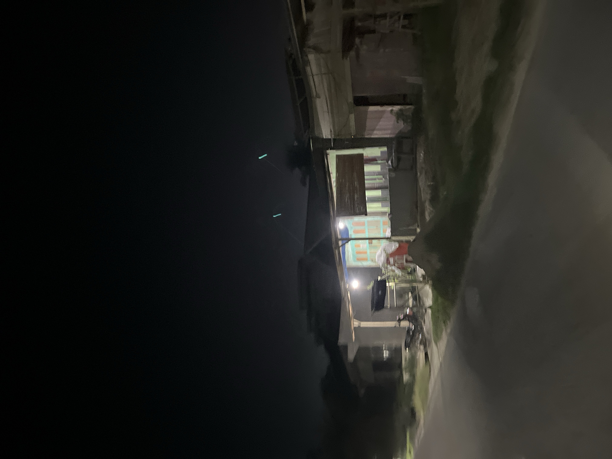 A slightly blurry, nighttime photograph captures a street-level view of a residential area in Bundar, Indonesia. The foreground shows a dark asphalt road. To the right of the road, there are two simple, open-fronted structures, possibly small shops or roadside stalls. One structure has a light-colored roof supported by posts and appears to be constructed with light-colored material. Inside this structure, there are some indeterminate objects and a red item, possibly a tarp or a piece of fabric. A motorcycle is parked on the ground in front of this structure. Adjacent to this, another structure is visible, appearing slightly more built, with what looks like a wooden door or shutters. A pile of dark material, resembling dirt or sand, is situated to the left of these structures. Further left, another building with a dark roof is partially visible. The background is dark, indicating a night scene, with faint lights suggesting the presence of other buildings or streetlights. Two faint, bluish-green lights are visible in the sky above the structures, which could be distant lights or possibly part of a drone or aircraft. The overall impression is one of a quiet, possibly rural or suburban, street scene at night.