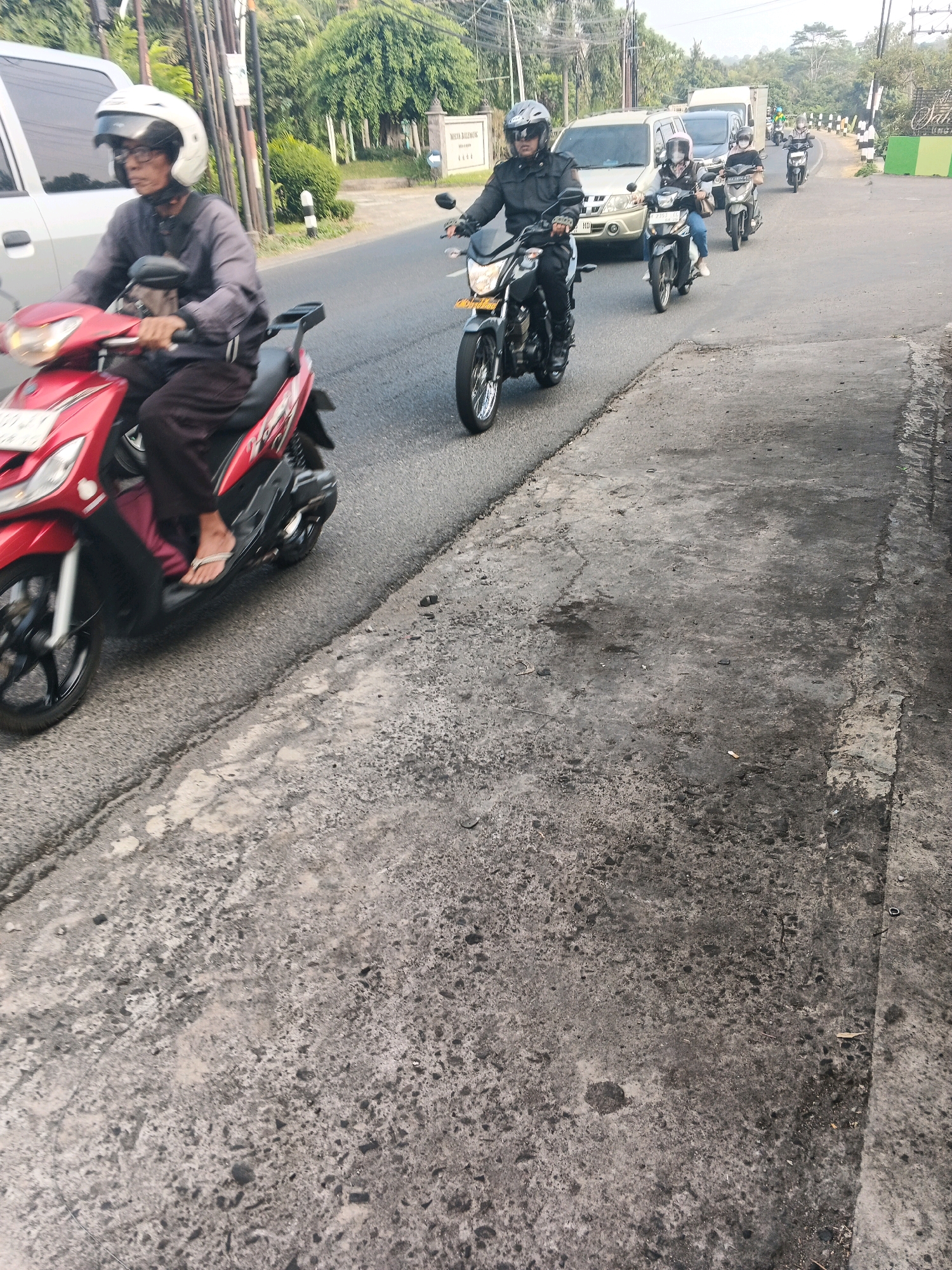 The image depicts a street scene in Ungaran, Indonesia, on a day with overcast skies, though the lighting suggests it is daytime. The foreground is dominated by a textured, grey asphalt road surface with visible patches of wear and debris.
In the mid-ground, a series of motorcycles and their riders are traveling along the road. Closest to the camera, a person wearing a white helmet and dark clothing rides a red and black scooter. Further down the road, another rider in a black helmet and jacket is on a black motorcycle. Behind them, several other motorcycles and riders are visible, including one with a passenger, all navigating the roadway. Some of the motorcycles have visible license plates.
The background reveals a street lined with trees and vegetation. A white van and a light-colored SUV are also present on the road. In the far background, a sign with Indonesian writing is visible, indicating a location or establishment. The overall impression is of moderate traffic on a local road in Ungaran.