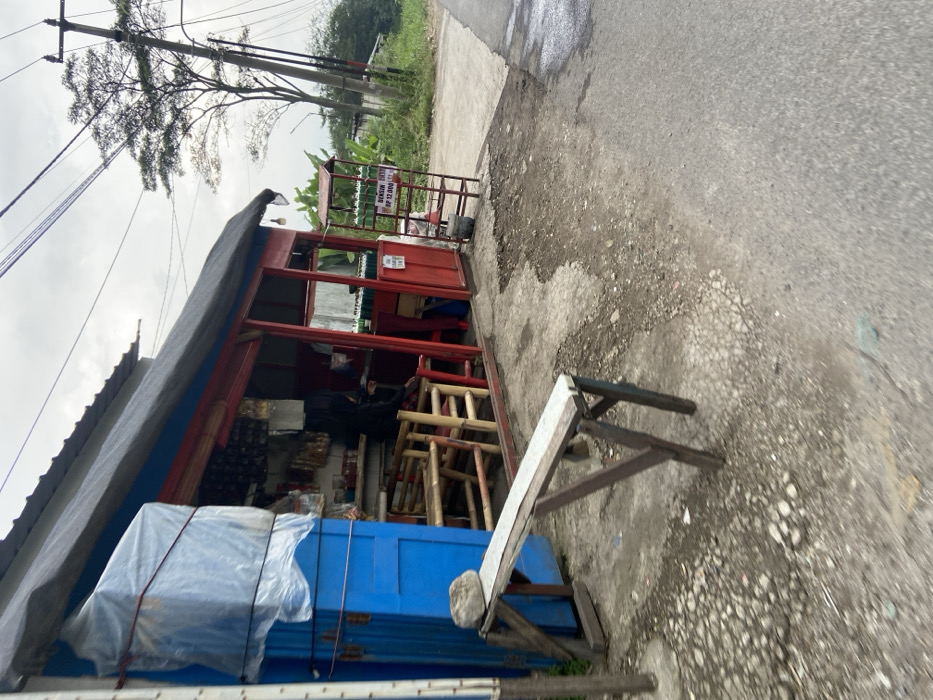 The image shows a small shop with a red painted exterior in Kecamatan Margaasih, Indonesia, located at coordinates (latitude: -6.963, longitude: 107.538). There is one person, partially obscured, seated in the shop. The shop has a blue painted door and is open to the front. There are several wooden chairs and tables in the shop. A wooden bench is sitting outside the shop. The shop is situated on a gravel path leading to a paved road with a tree line on the right side. The shop has a small metal shelf displaying goods for sale. The shelf has a sign with the words "Bensin Pertalite" written on it. The area is partially obscured by a power line, which is supported by a wooden pole. There are numerous power lines visible in the sky. The sky is a light gray and overcast. The road appears to be in need of repairs, with many potholes visible.