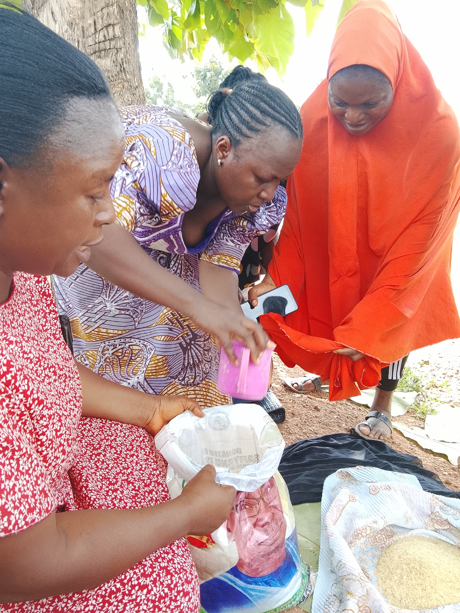 Three women are gathered outdoors, possibly in a market or on a roadside, near a tree. The woman on the left, wearing a red and white patterned dress, is holding an open bag containing a white granular substance, possibly rice. Her head is tilted down as she focuses on the contents of the bag.
The woman in the middle, wearing a purple and gold patterned dress, is leaning over and appears to be pouring something from a pink container into the bag held by the woman on the left. She is looking down at her hands.
The woman on the right is wearing an orange headscarf and a matching orange garment. She is holding a portion of her orange garment and looking down, as if inspecting it or handing it to someone. A smartphone is visible in her hand, reflecting the image of a person's face.
The ground is covered with dirt and some loose fabric. In the background, there are green leaves from a tree and a bright sky, suggesting it is daytime and likely sunny. The overall scene depicts a moment of commerce or sharing, with the women actively engaged with the items in front of them. There is no visible text in the image.