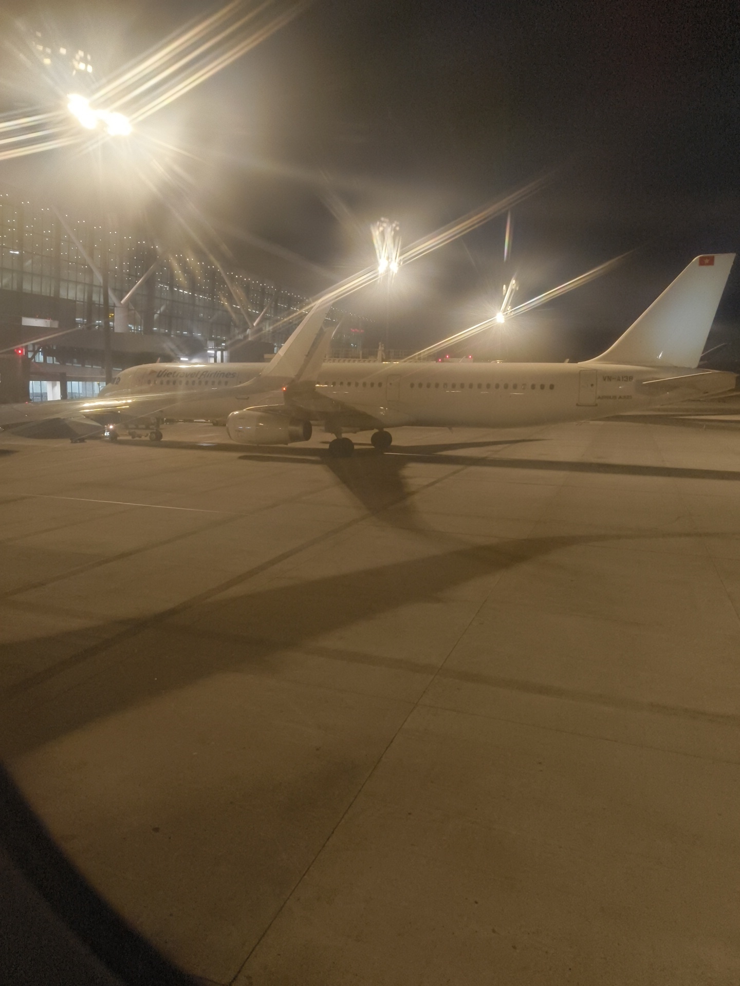 An Airbus A321 aircraft with registration VN-A138, operated by Vietravel Airlines, is parked at an airport gate at night. The aircraft is predominantly white with the airline's branding visible on the fuselage. Bright lights illuminate the scene, casting long shadows from the aircraft onto the tarmac. The background shows a modern airport terminal building with large windows and internal lighting. The image appears to have been taken in Thuận An, Vietnam, likely at Tân Sơn Nhất International Airport, given the airline and typical aircraft operations in that region.