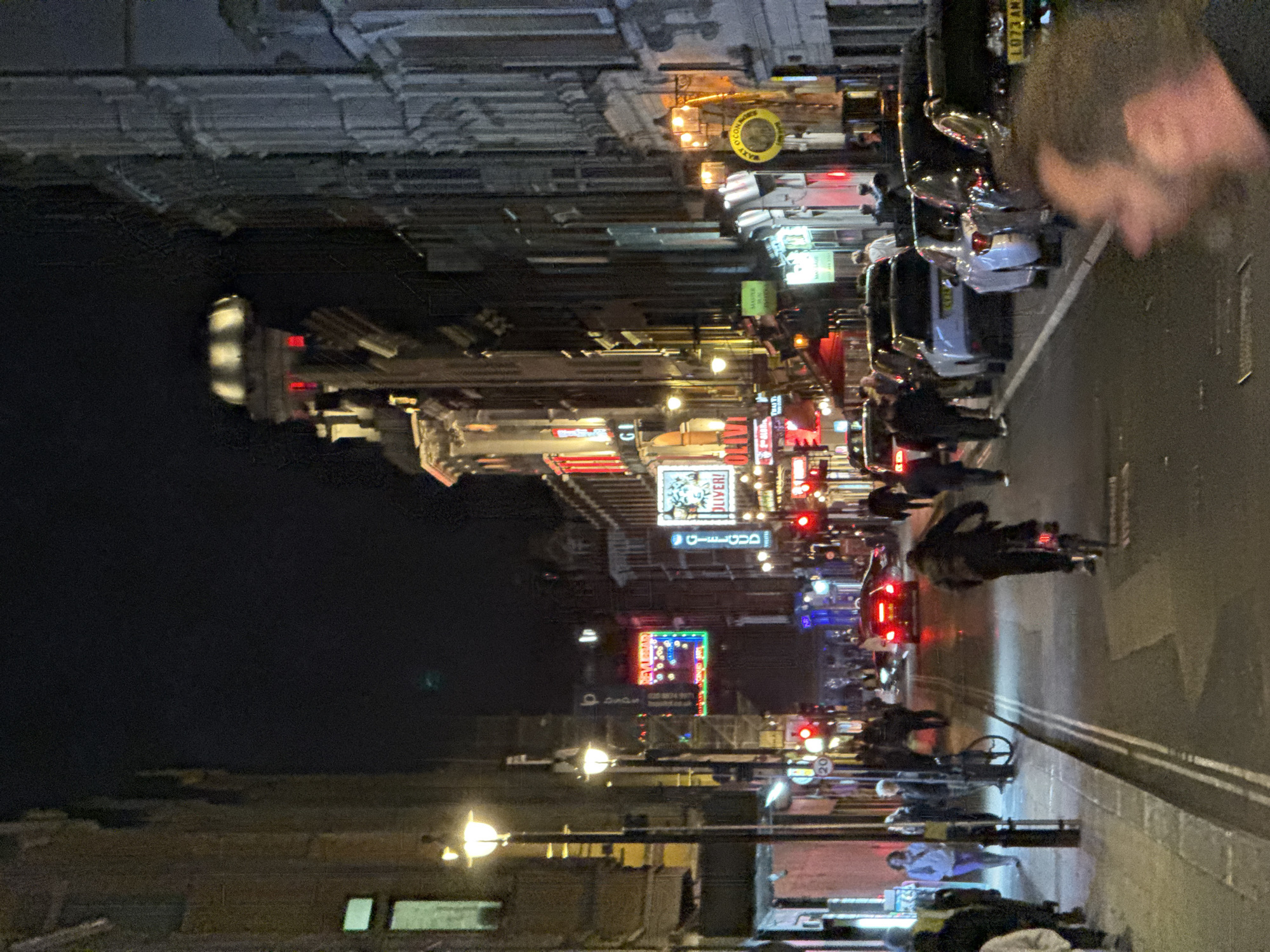 This nighttime street scene shows a bustling street in London, United Kingdom. The camera angle is rotated to the left. People on foot and bicycles share the road with cars, their lights reflecting on the wet pavement. Buildings line the street, illuminated by various signs and streetlights. Signage visible includes "Deliver!" and Arabic script. The street is lined with parked cars. A person's face appears partially in the upper right corner of the frame. The overall impression is of a vibrant urban environment at night.
