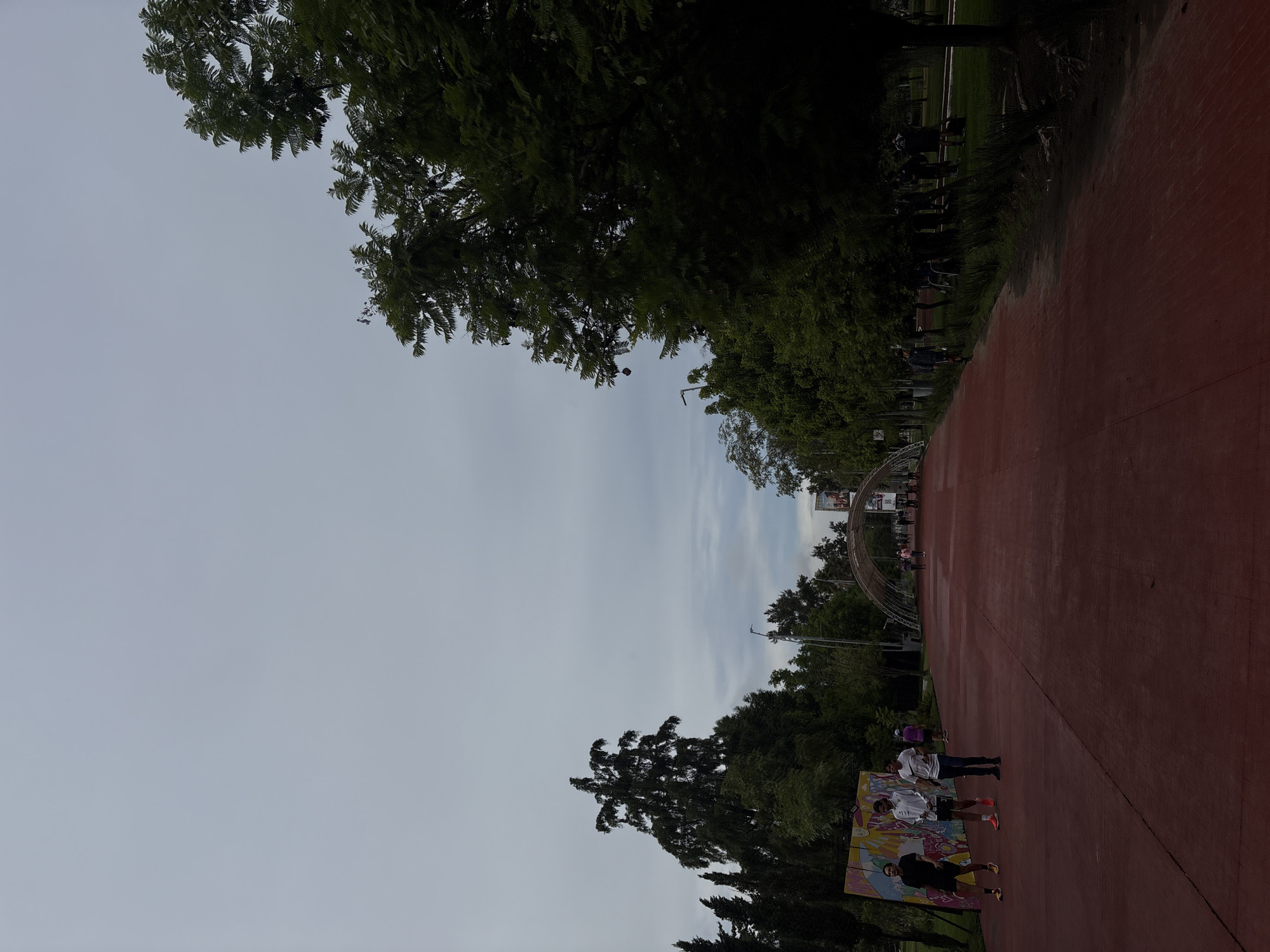 The image depicts a wide, red-colored path, likely a jogging or biking track, in a park. The path stretches into the distance and is bordered by lush green trees on both sides. In the background, an arched structure, possibly an entrance or a decorative element, spans the path. Several individuals are visible on the path, some walking and others appearing to be jogging. On the left side of the path, a colorful mural is mounted on a large panel. The sky is overcast, suggesting a cloudy or potentially rainy day. The overall atmosphere is one of an outdoor recreational space, possibly a park in a city. Based on the presence of similar arched structures and vibrant murals often found in such settings, this location could be the 'Parque Lineal' or Linear Park in Puebla, Mexico.