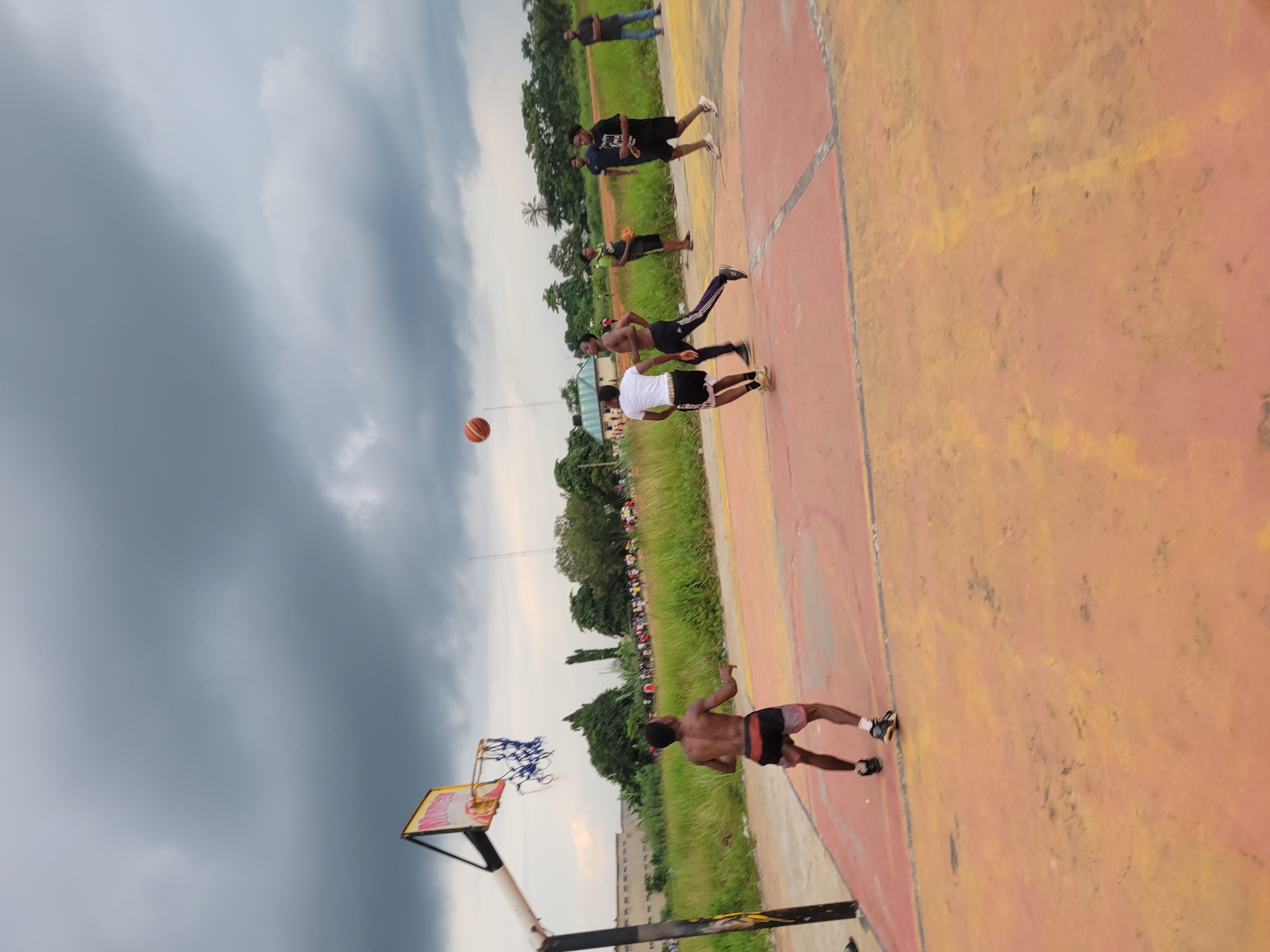 The image depicts an outdoor basketball court in Egbagu, Nigeria, under a cloudy sky. Four young men are engaged in a basketball game. One player in the foreground, wearing black shorts and white socks, is positioned on the court with his back to the camera, seemingly focused on the game. Another player, wearing black shorts and a white t-shirt, is in motion, facing away from the camera and towards the basket. To his right, a third player, bare-chested with black shorts, is running with the ball. In the background, two other men are observing the game; one in a black t-shirt and shorts, and another further to the right in a black t-shirt and blue jeans. The court itself is a faded orange and yellow surface, marked with black lines. Behind the court, a grassy field extends to a treeline, with some buildings visible in the distance. A basketball hoop with a frayed net stands on the left side of the court. The overall atmosphere suggests a casual game in progress.