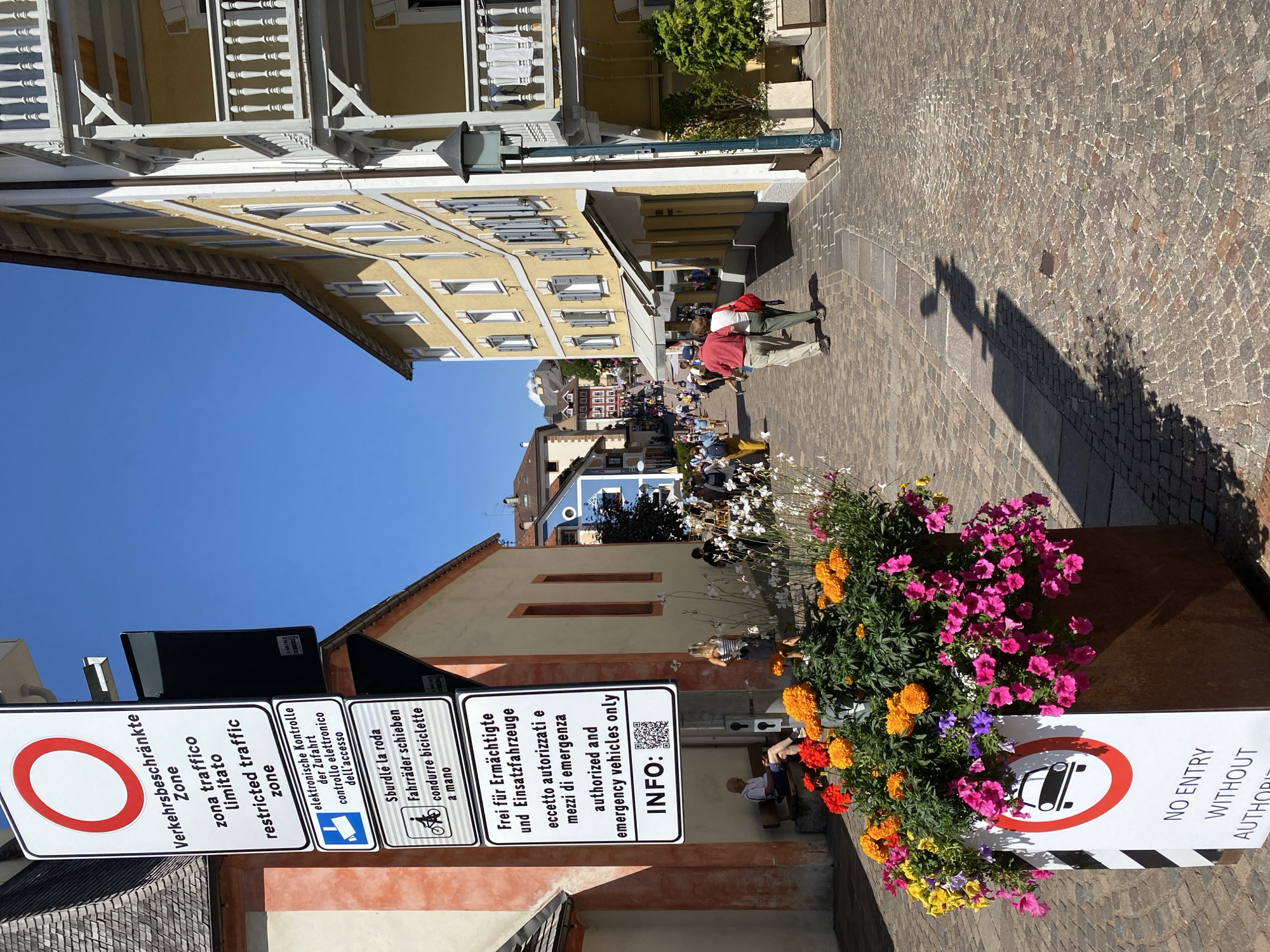 The image depicts a sunlit street scene in Urtijëi - St. Ulrich in Gröden - Ortisei, Italy. A tall, multi-panel signpost stands on the left, displaying various traffic regulations in multiple languages, including a "Restricted Traffic Zone" symbol. Below these signs, a large planter filled with colorful flowers, predominantly pink and orange, sits in the foreground, with a smaller "No Entry Without Authority" sign adorned with a car symbol.
The street is paved with cobblestones, and the buildings lining it feature typical alpine architecture with light yellow facades and wooden balconies. Several people are walking along the street, some individually and others in small groups. On the left, a man sits on a bench, looking at his phone. On the right, a couple walks away from the camera. In the background, more people are visible walking further up the street, suggesting a lively atmosphere. The sky is clear and blue, indicating fair weather. The overall impression is of a charming and busy pedestrian area in a picturesque Italian town.