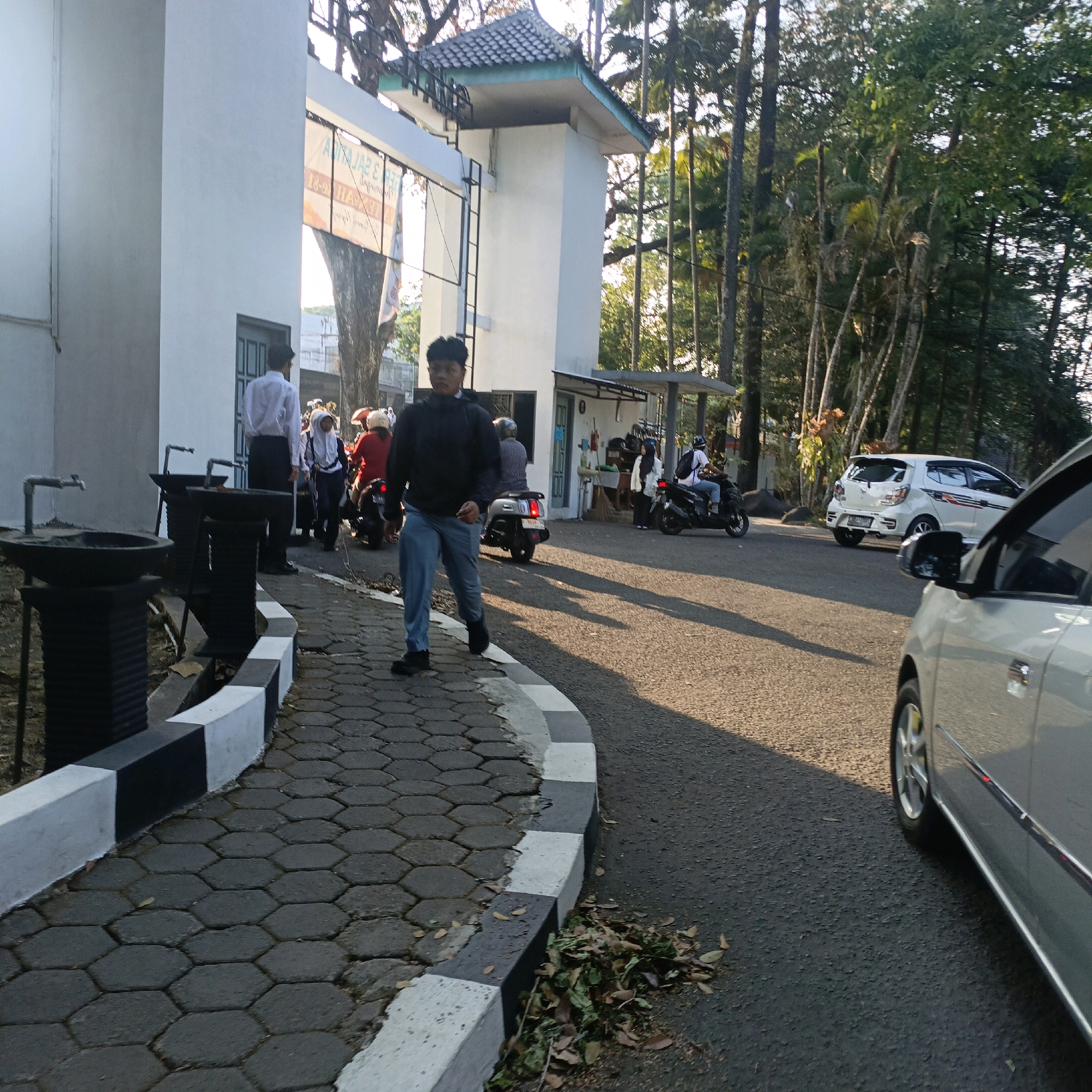 The image depicts an outdoor scene, likely on the grounds of an educational institution in Salatiga, Indonesia, given the presence of students and uniformed individuals. In the foreground, a person in a black jacket and blue jeans walks on a hexagonal-tiled path bordered by a curb painted with alternating black and white stripes. To the left, there are black rectangular planters with faucets.
Further into the scene, a white car with red and black decals is parked on the asphalt road. Several individuals are present, some on motorcycles. One person in a white hijab and blue pants walks towards the road. Behind them, another person is seated on a motorcycle. More people are visible near a building with a white facade and a dark-tiled roof, suggesting an entrance or administrative building. Tall trees with lush green foliage line the background, indicating a possibly tropical or sub-tropical environment. The lighting suggests it is daytime, with sunlight casting shadows on the road.