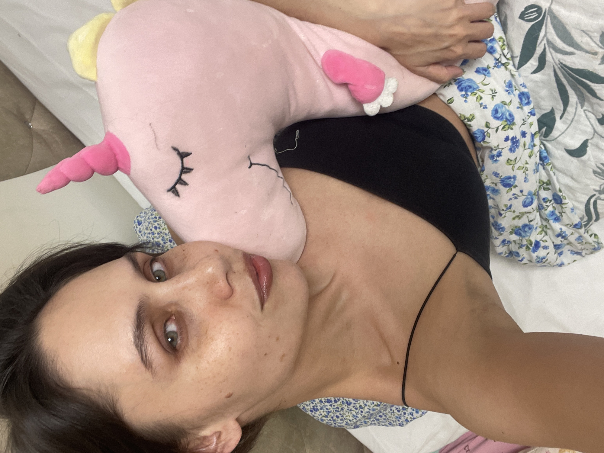 A woman lies in bed, holding a pink unicorn stuffed animal. She is wearing a black top and is partially covered by a floral-patterned blanket. The location is unspecified and there is no information suggesting that this image was taken in Izhevsk, Russia.