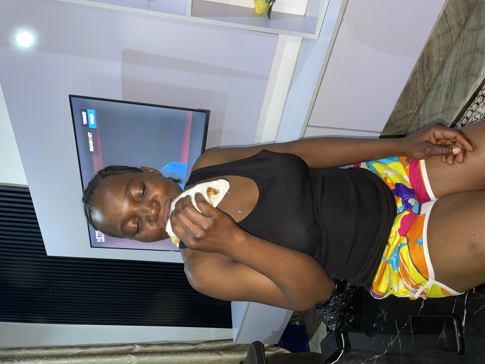 A woman is seated indoors. She is eating a food item wrapped in a white napkin or paper. She wears a black tank top and colorful shorts. Behind her, there is a television mounted on a white wall, and a white shelf is above it. The exact location cannot be determined from the image alone, but it is speculated to be Jos, Nigeria.