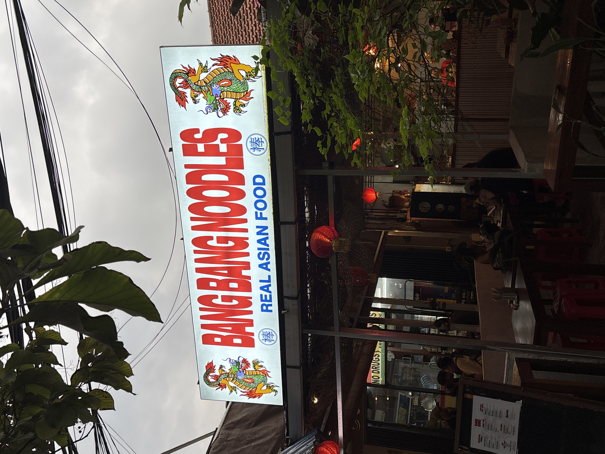 The image displays the brightly lit vertical sign of "BANG BANG NOODLES," an Asian restaurant in North Kuta, Indonesia, likely captured at dusk or early evening. The sign features the restaurant name in large red letters, with "REAL ASIAN FOOD" in blue below it, flanked by two Chinese characters (棒), and adorned with colorful dragon illustrations. A cloudy sky forms the backdrop, crisscrossed by numerous black overhead electrical wires. In the foreground, large green leaves partially obscure the view.
The restaurant's entrance is decorated with traditional red lanterns, and lush green foliage hangs around the structure, contributing to a tropical ambiance. Inside, several patrons are seated at tables, suggesting dining activity, though details of their emotions are not discernible. One person inside appears to be wearing a helmet. A smaller sign or sticker visible on a window panel reads "NO DRUGS," along with partial text including "FR," "SE," "PE NO.1," and "UNIA." The overall scene depicts a casual, open-air dining establishment common in the region.