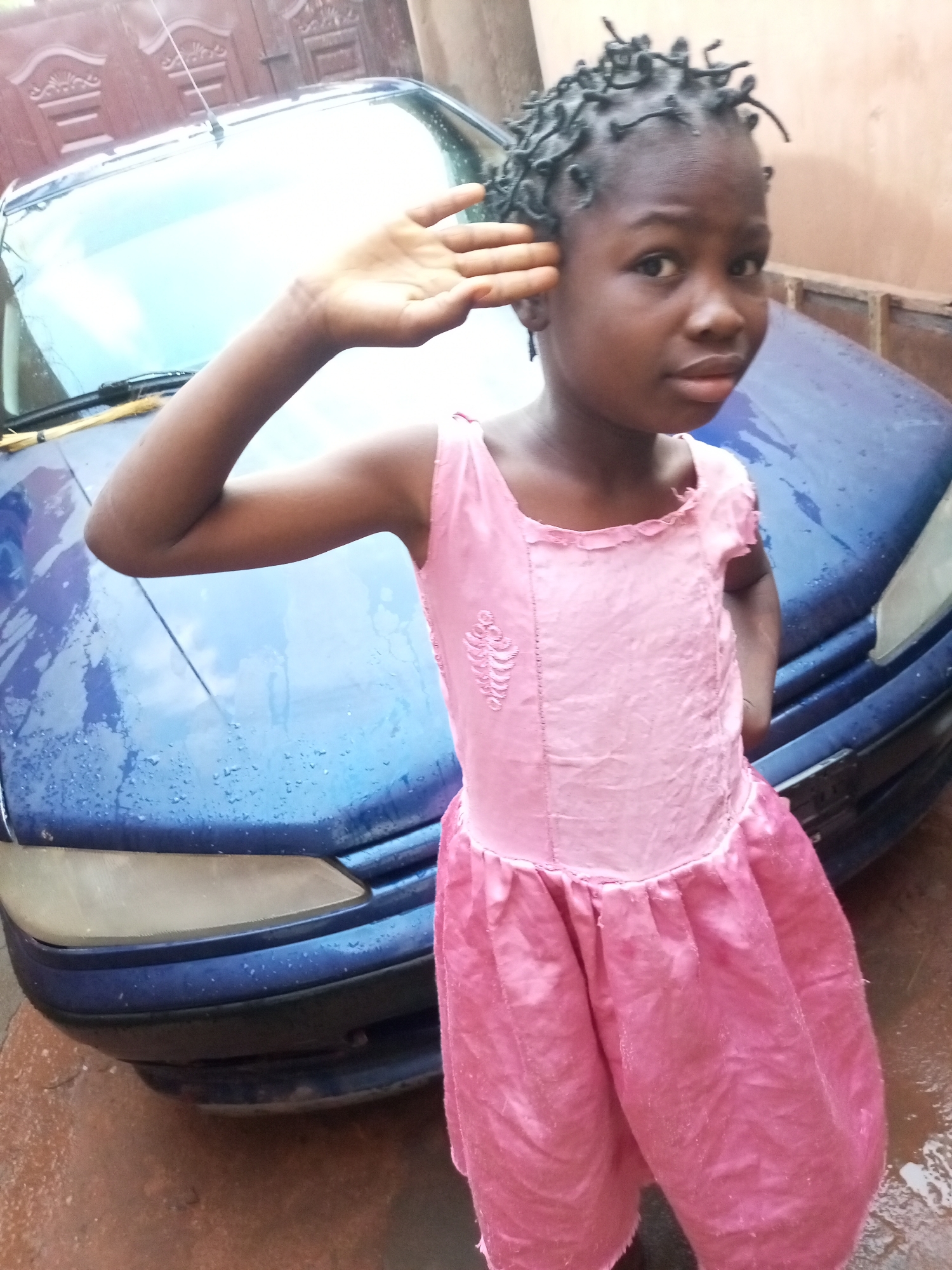 A young Black girl with dark skin and braided hair stands in front of a blue car. She is wearing a pink dress with a ruffled neckline and sleeves, and a gathered skirt. Her right arm is raised to her forehead in a saluting gesture. The car is wet, suggesting it may have recently rained or been washed. The background appears to be a residential area with a reddish-brown wall and a patterned gate. The ground is also wet and appears to be paved with a rough texture.
