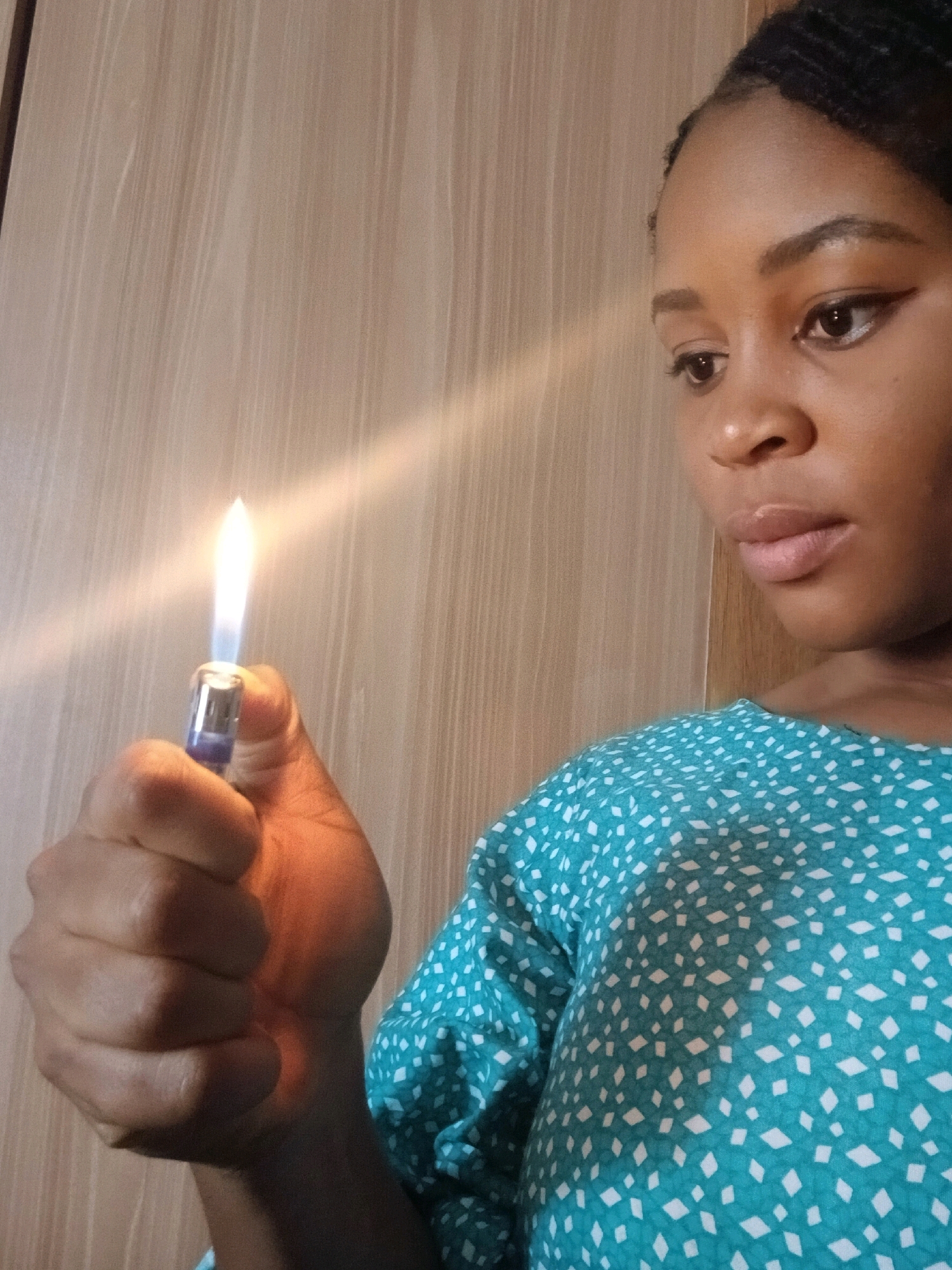 In Gura Top, Nigeria, a close-up shot shows a young Black woman in the foreground, holding a lit blue and silver lighter in her right hand. The flame emits a bright light and casts a warm glow on her skin. She is wearing a teal colored shirt with a white diamond pattern. Her facial features are visible, including her eyebrows, eyes, nose, and lips. Her hair appears to be styled in braids. The background consists of a wooden surface, possibly a closet door, with vertical grain lines. The lighting suggests an indoor setting with a light source coming from the left.