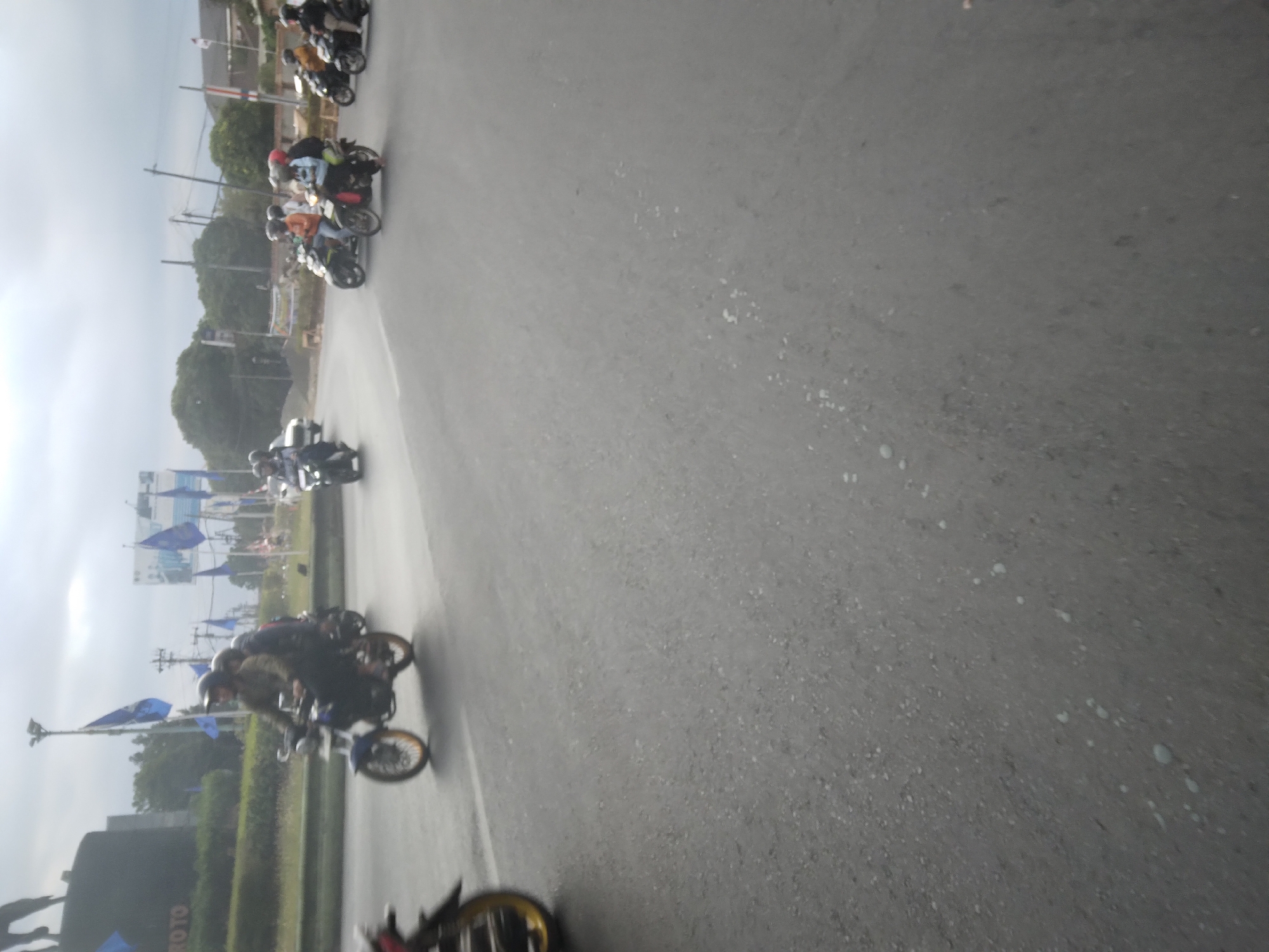 The image captures a street scene in Desa Pamijen, Kecamatan Sokaraja, Indonesia, with several motorcycles and riders. In the foreground, a motorcycle with a rider and a passenger is visible. The rider is wearing a helmet and a brown jacket, while the passenger appears to be wearing a dark top. To the left, a portion of another motorcycle with a yellow rim is partially visible. In the background, multiple other motorcycles are seen, some with multiple passengers, navigating the asphalt road. The environment appears to be urban, with trees and poles lining the road, and flags are prominently displayed in the background. The sky is overcast.