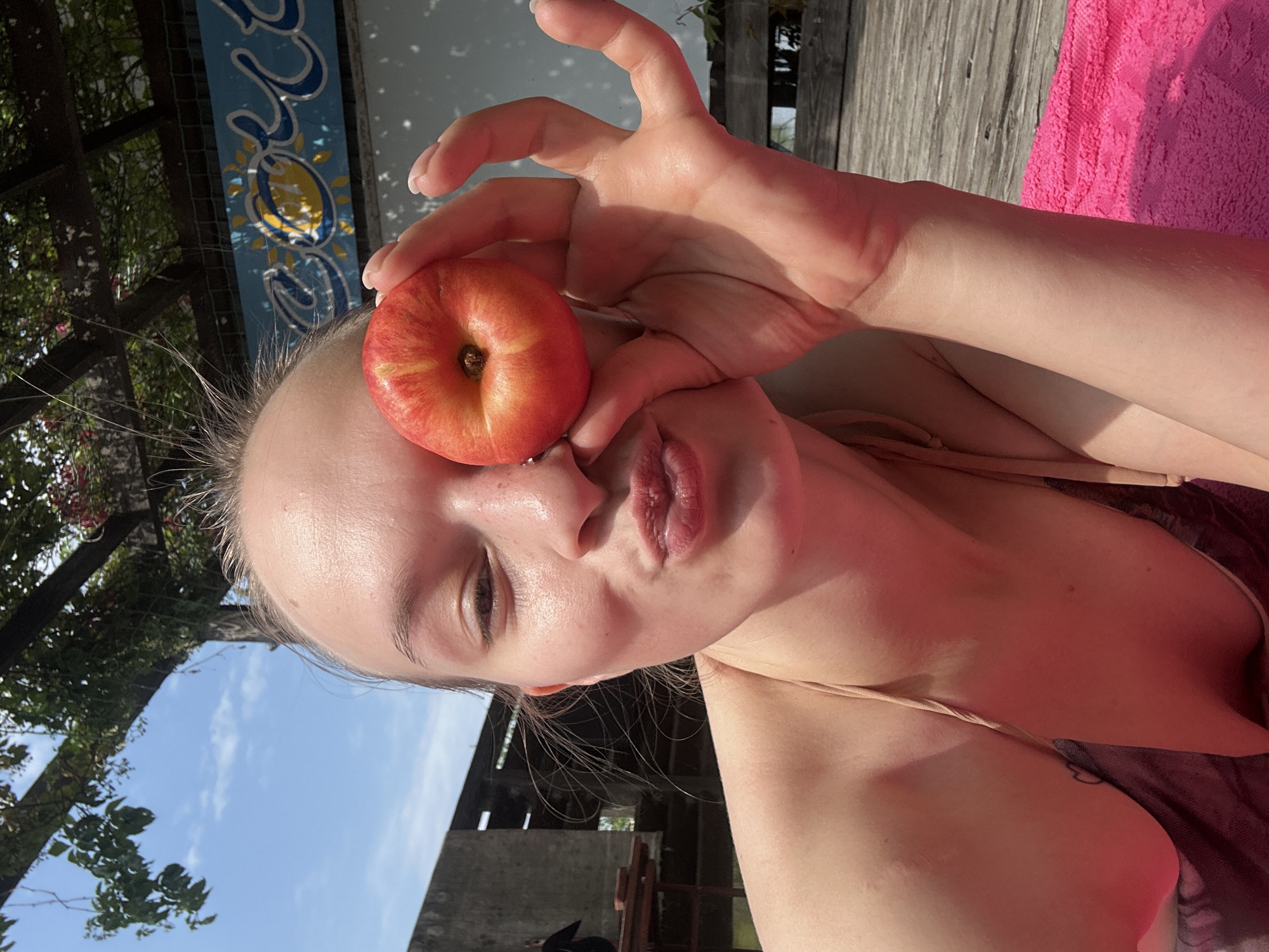 A young woman with fair skin and blonde hair pulled back is holding a red and yellow apple in front of her right eye. She is wearing a light-colored bikini top and has her lips pursed. Her left hand is visible, with fingers extended. She is outdoors, with a blue sky and greenery in the background. A wooden deck and a pink towel are in the foreground. There is a building with a sign that reads "COCO" behind her. The image does not contain any information indicating the location is in Chaikovsky, Russia.
