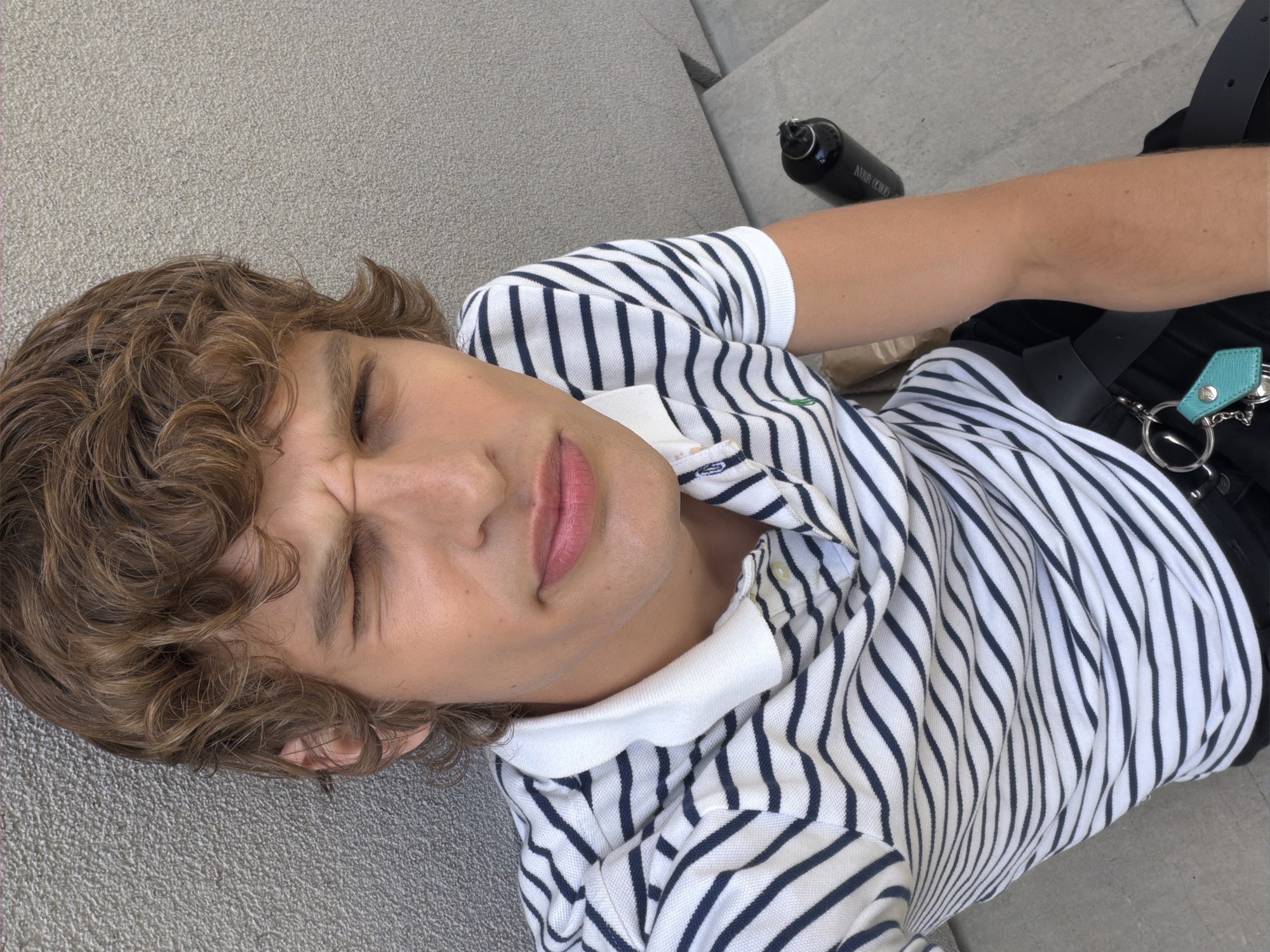 A young person with light brown, curly hair is depicted from a high angle, lying on a light grey, textured surface, possibly concrete or stone steps, in what appears to be an outdoor urban setting in Milan, Italy. Their eyes are closed or heavily squinting, and their face is slightly scrunched, which could indicate a strong light source or a deliberate, playful expression.
They are wearing a casual polo shirt with thin navy blue horizontal stripes on a white background, featuring a white collar. A small green embroidered logo, resembling a polo pony, is visible on the upper left chest area of the shirt. To their right, a black water bottle is placed on the grey surface; the word "ARMANI" is clearly visible in white text on its side. Part of a black bag or harness and a teal-colored keyring or charm attached to a metal ring are also visible. The ambient lighting suggests it is daytime.