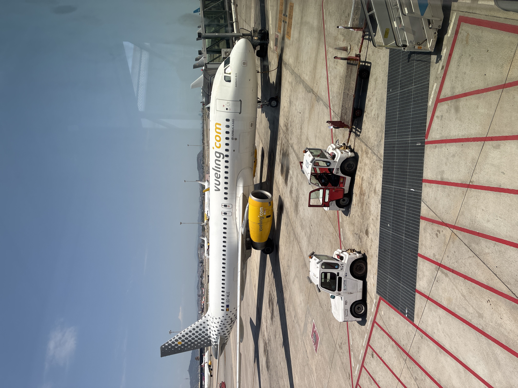 The image shows an airplane at an airport gate on a sunny day. The airplane is white with a grey and yellow pattern on its tail and yellow engines. The airline name "vueling.com" is visible on the side of the plane. In front of the plane are two small airport vehicles, a white tug and a red and white tug, and a baggage cart. The airport tarmac is marked with red and white lines. In the background, other airplanes and airport structures can be seen. The scene is observed through a window, with reflections visible on the glass. The location is not Großbeeren, Germany.